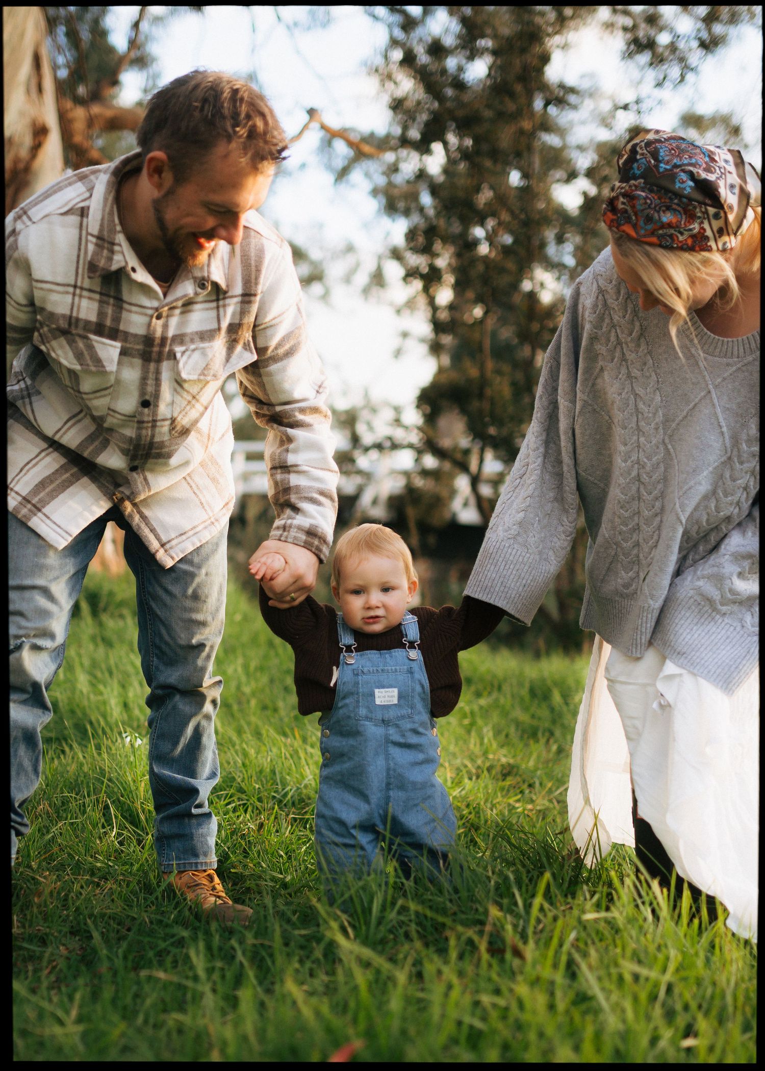 Parents helping toddler in denim overalls take first steps on green grass outdoors.