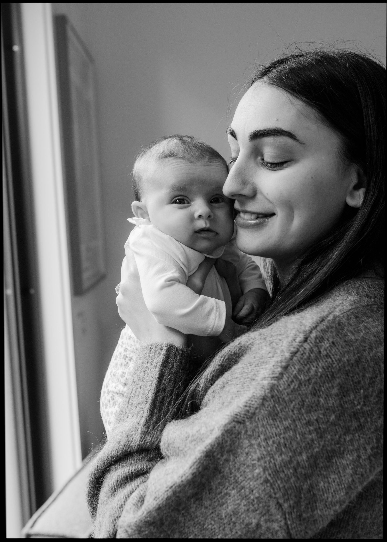 Black and white portrait of smiling mother holding alert newborn baby close to her cheek.