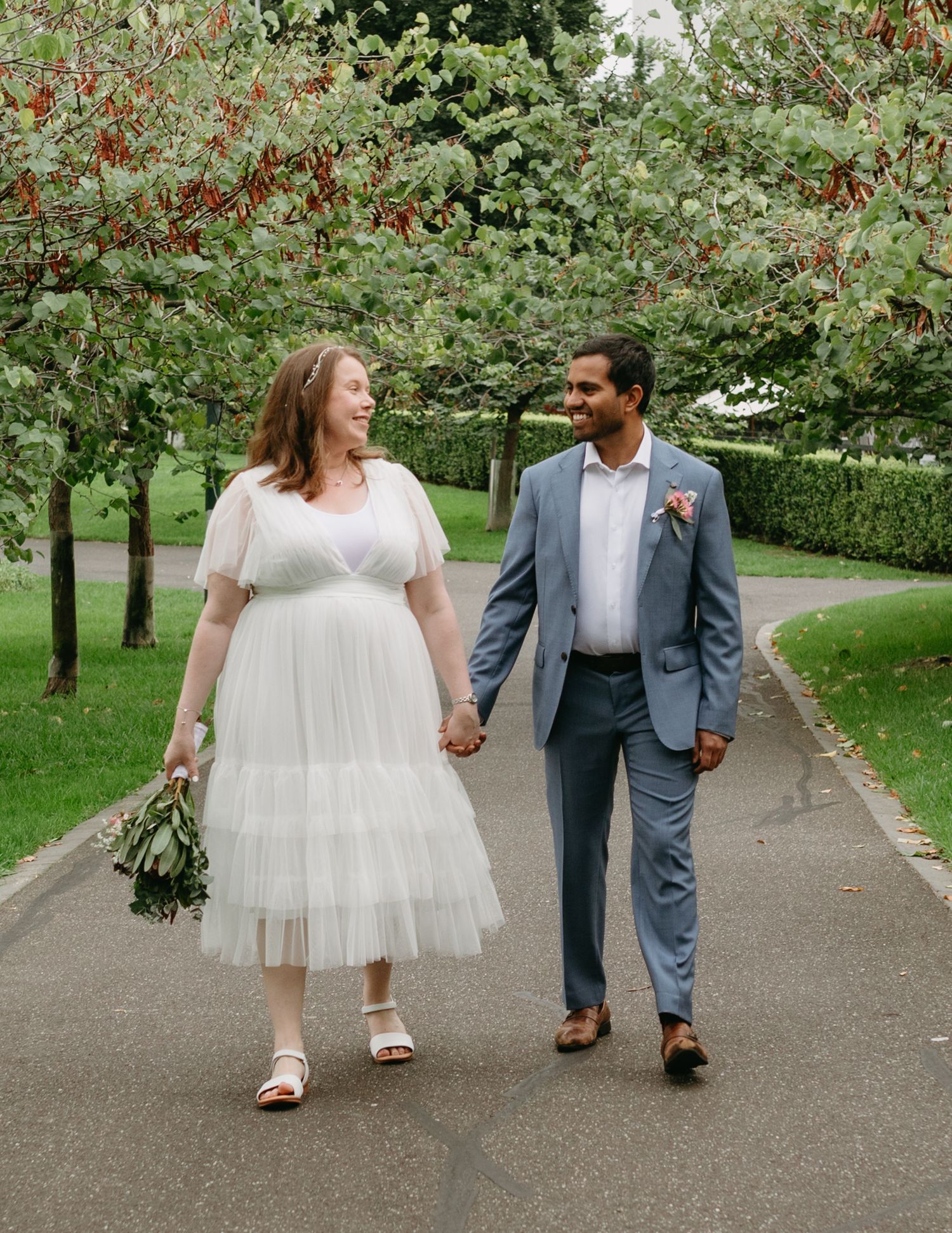 Bride and groom walking hand in hand through a tree-lined park path, bride holding a green bouquet.
