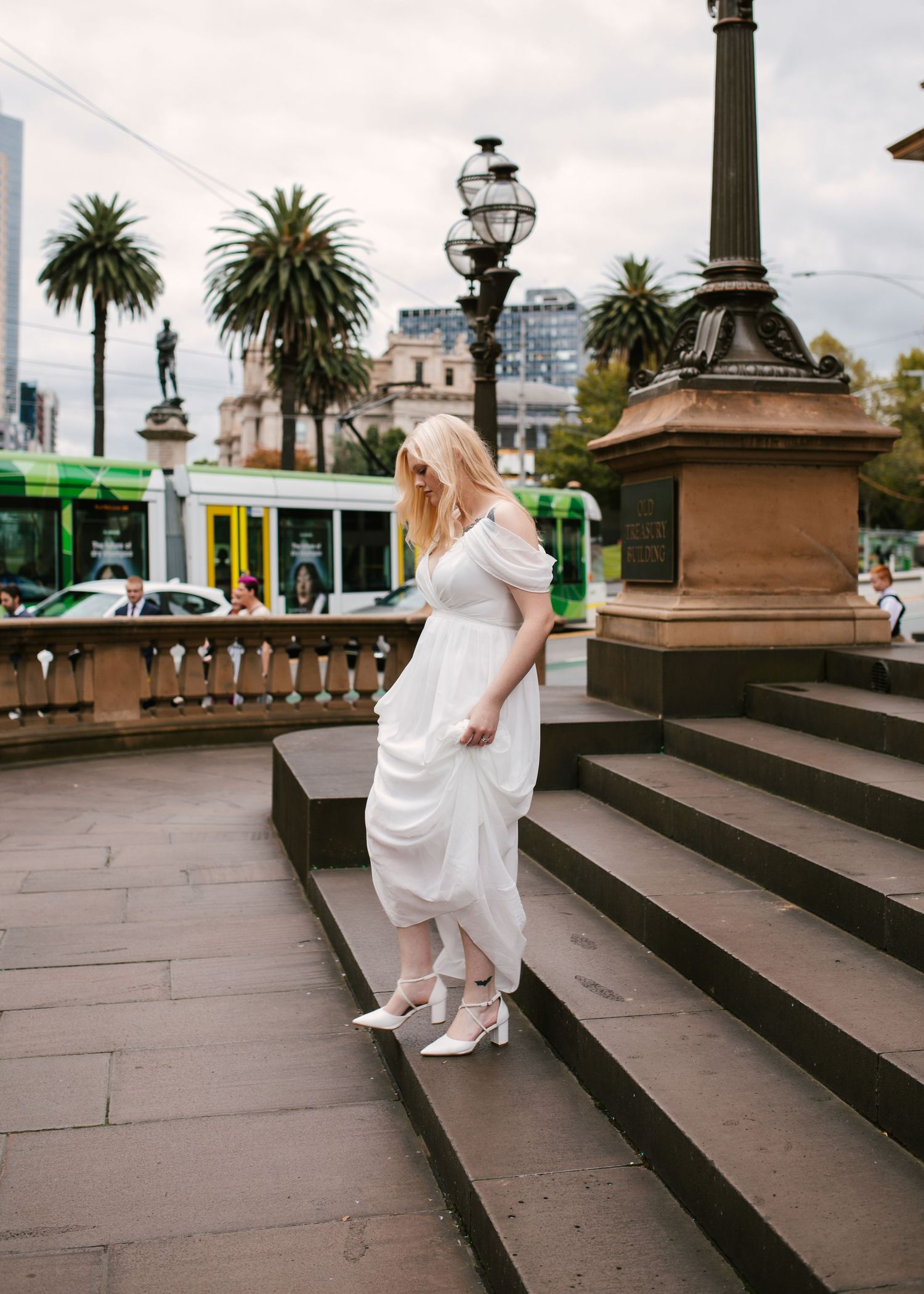 Blonde bride in white off-shoulder dress and heels descending stone steps near Melbourne tram stop.