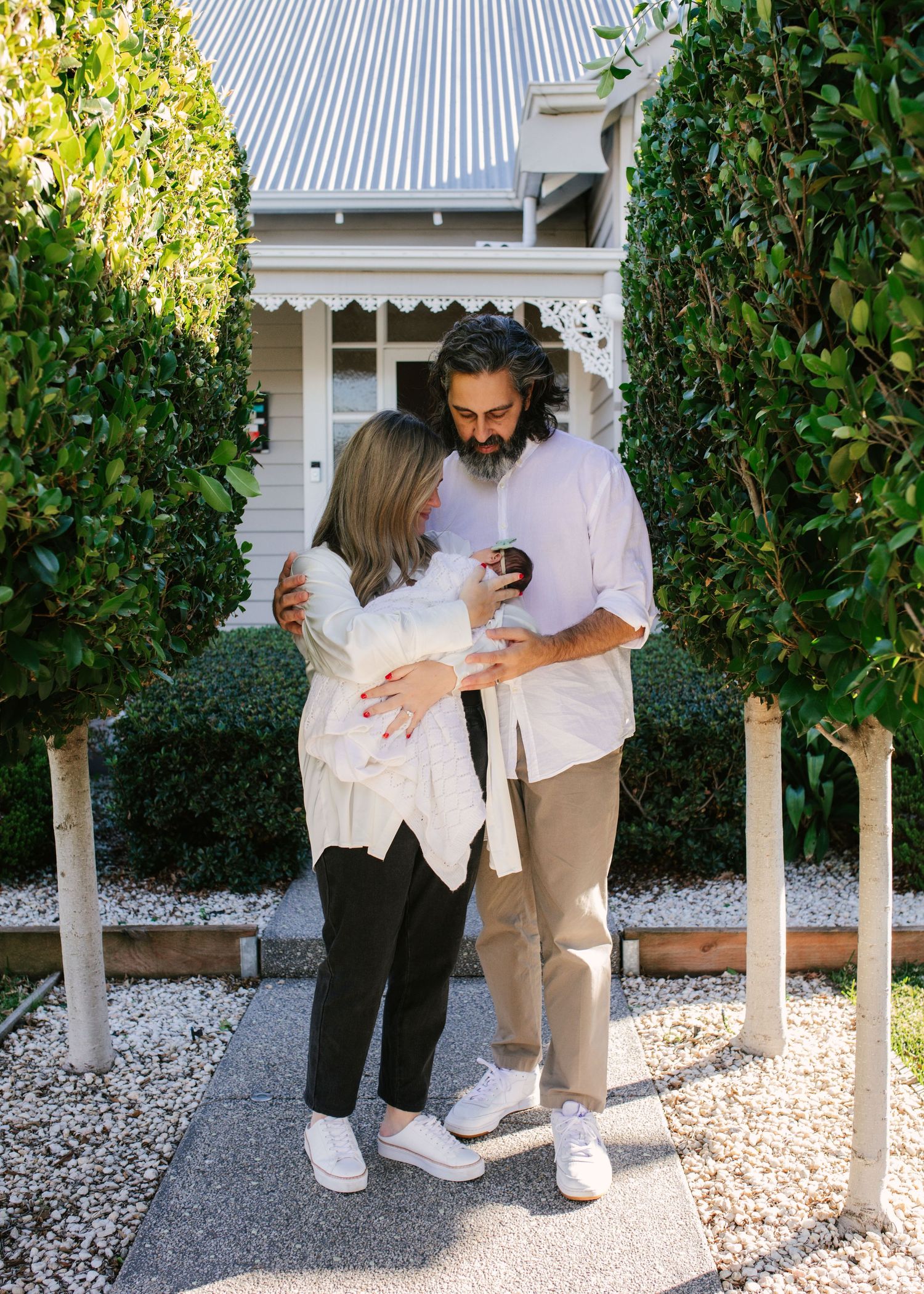 Happy couple standing in garden pathway holding newborn baby outside their charming cottage home.