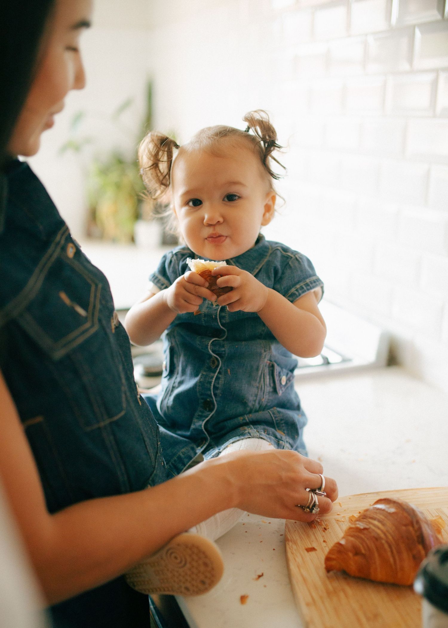Adorable toddler girl in denim dress eating a croissant while sitting on a kitchen counter.