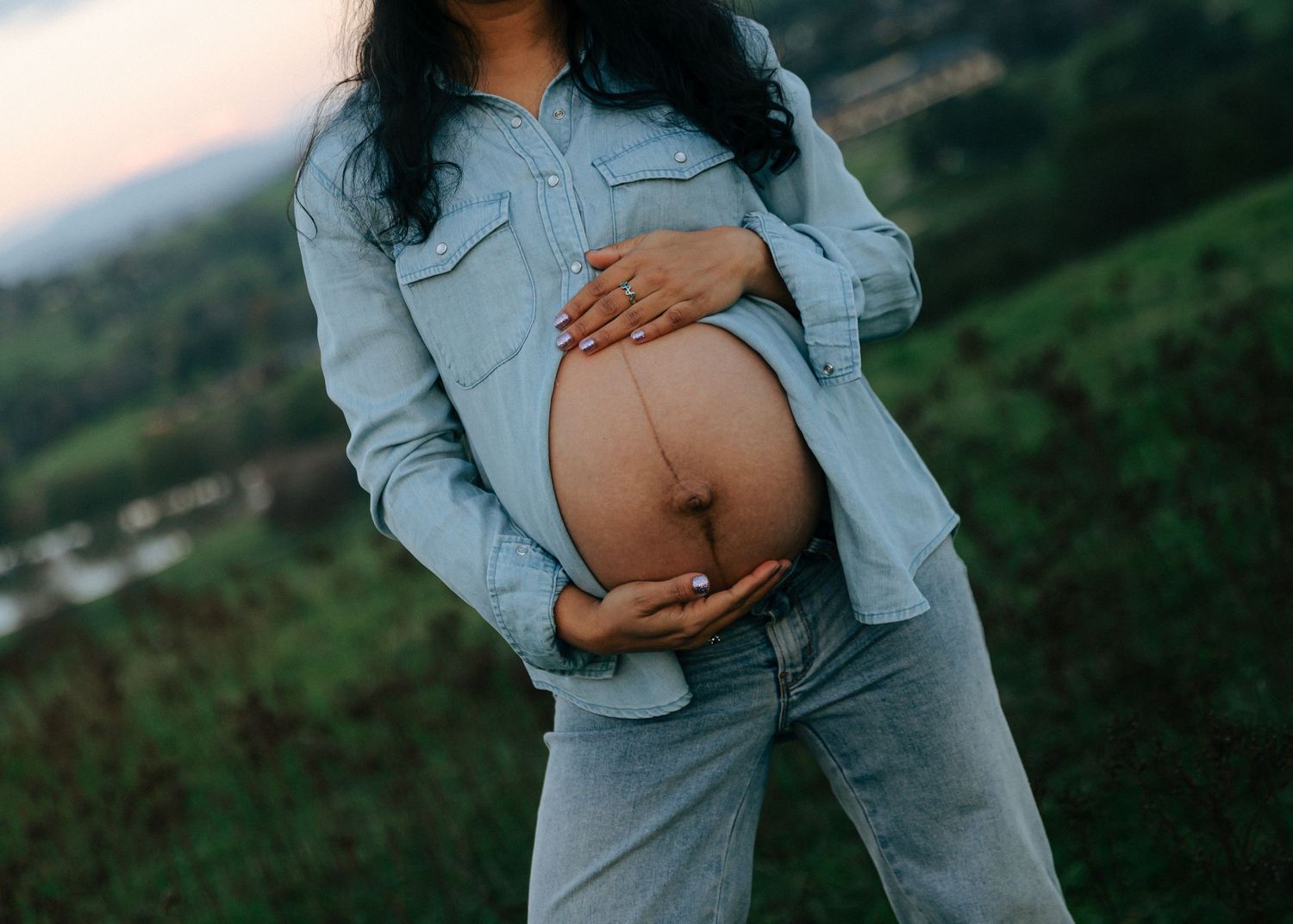 Pregnant woman in denim outfit cradling bare baby bump outdoors with green hills in background.