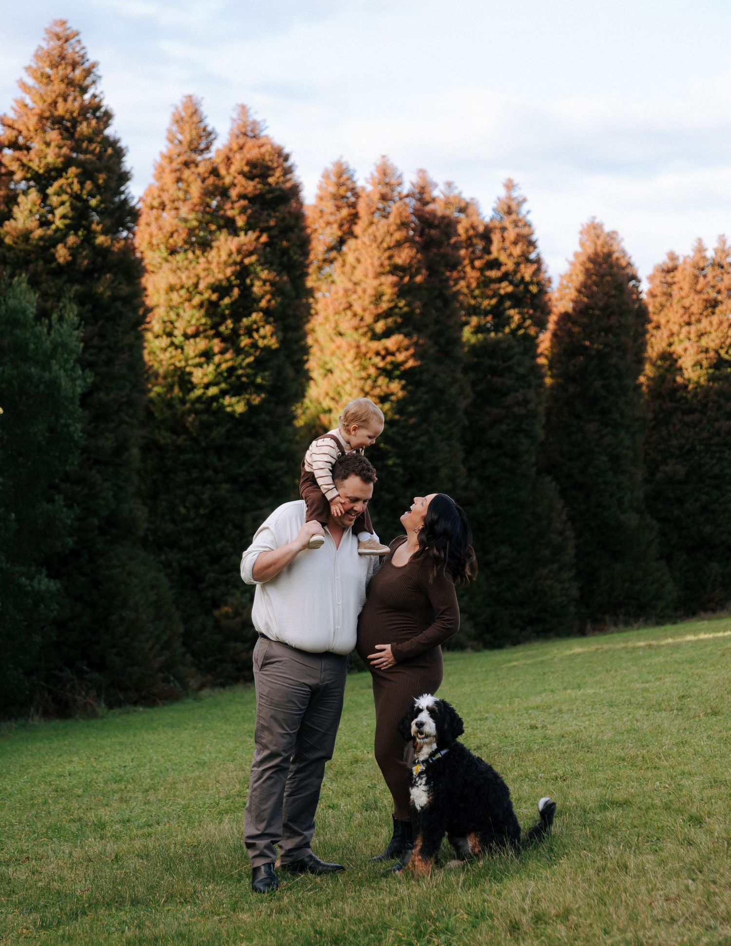 Pregnant woman in brown dress with husband, toddler, and Bernese Mountain Dog in autumn garden.