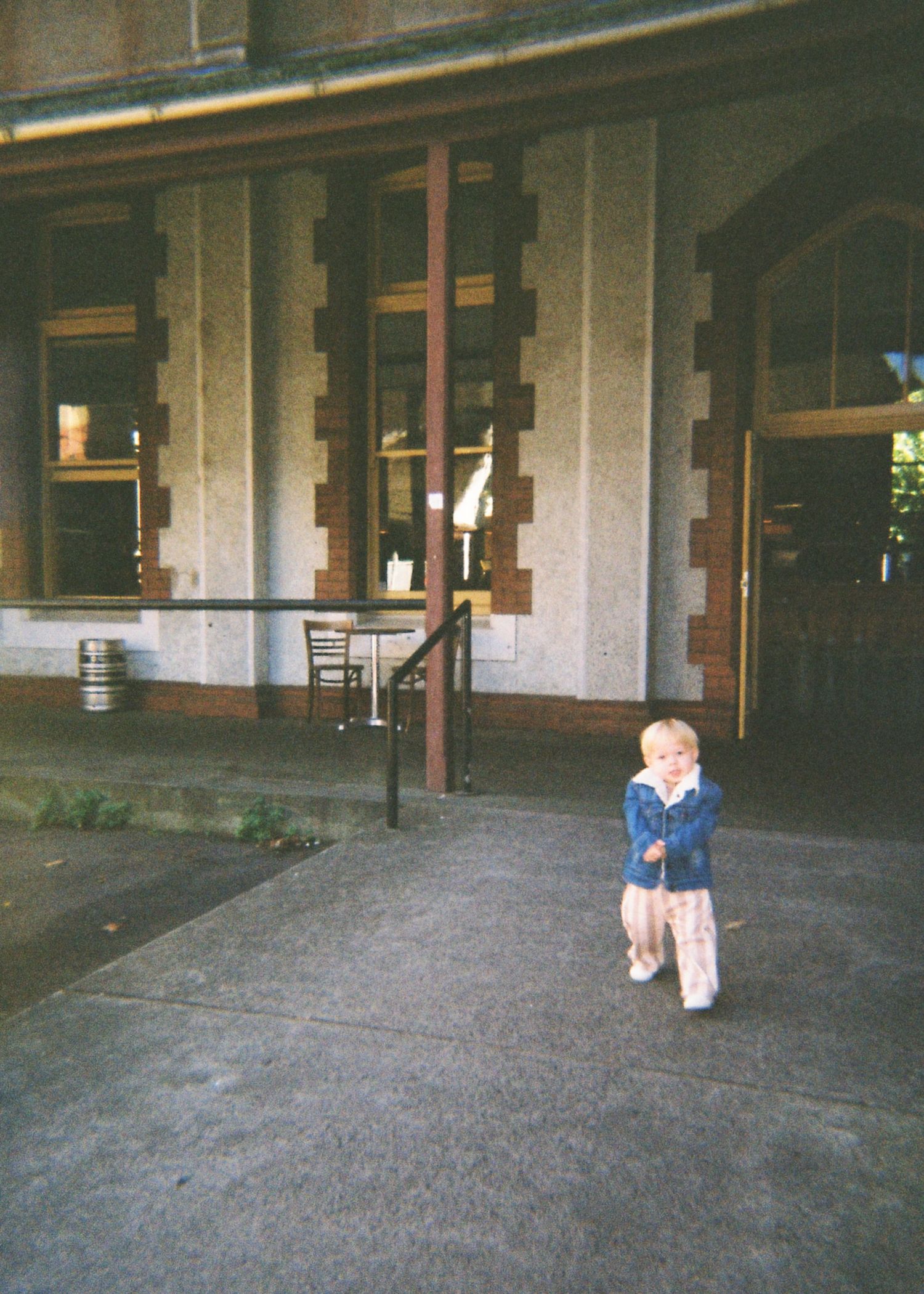 A young toddler walks along a sidewalk in front of a historic stone building entrance.