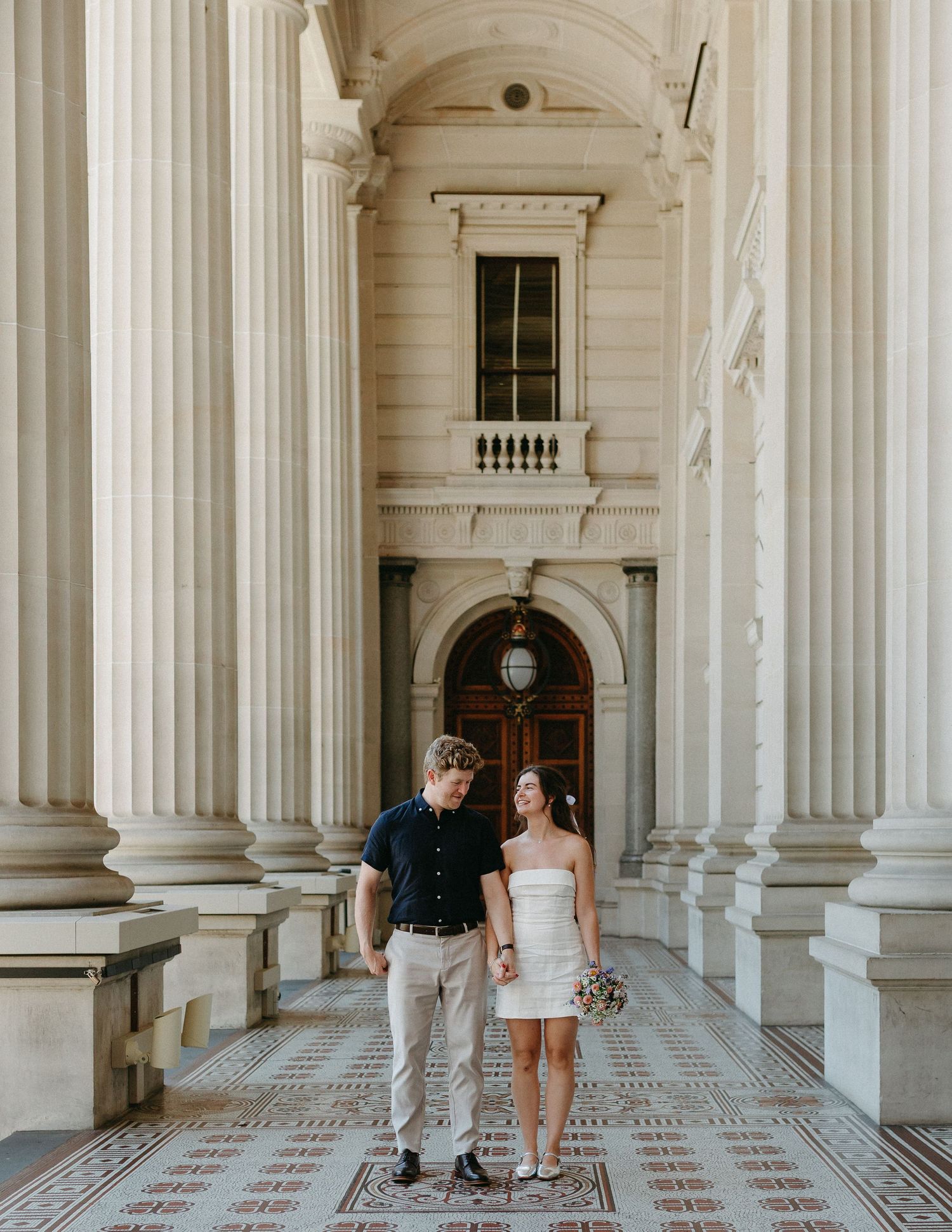 A couple holds hands and smiles while walking through grand white marble columns of a stately building.