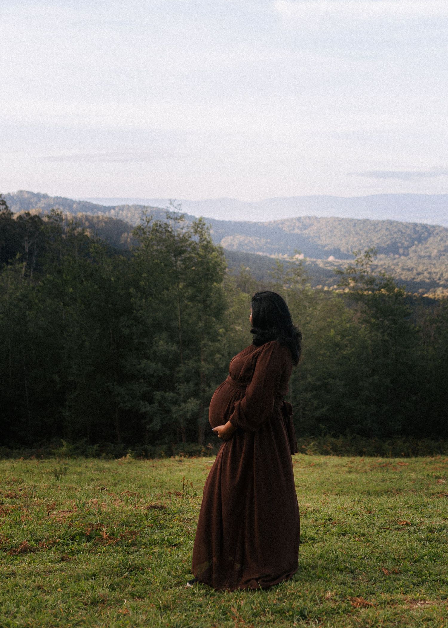 Pregnant woman in a flowing brown dress cradles her belly while standing on a grassy hilltop overlooking forested mountains.