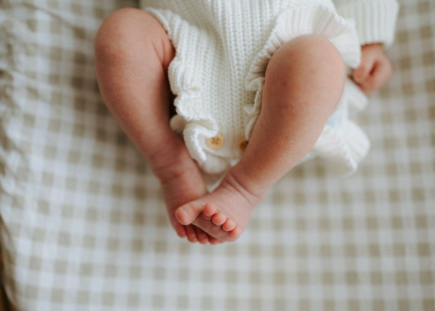 Overhead view of newborn baby tiny feet and legs in white knit outfit on neutral gingham fabric.