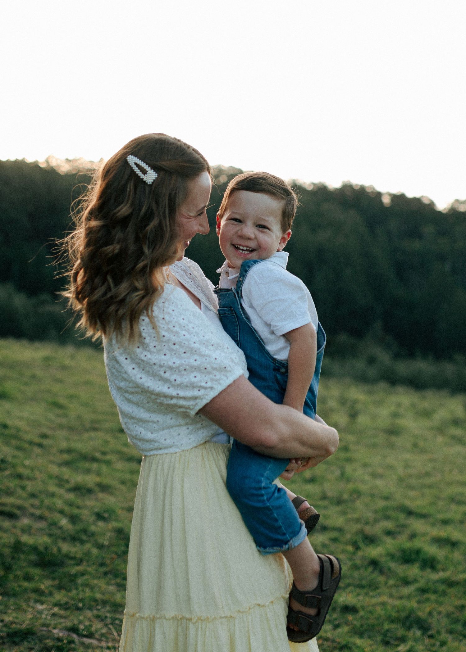 A smiling mother holds her cheerful toddler son in a golden hour outdoor field portrait.