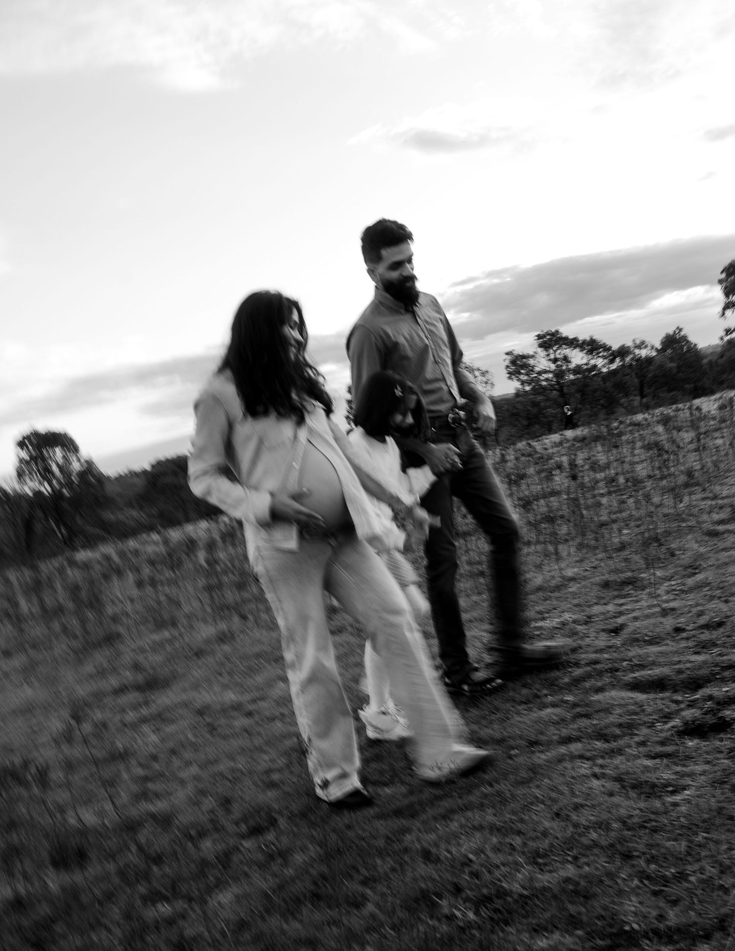 Black and white photo of pregnant woman and partner walking through a field at dusk in motion blur.