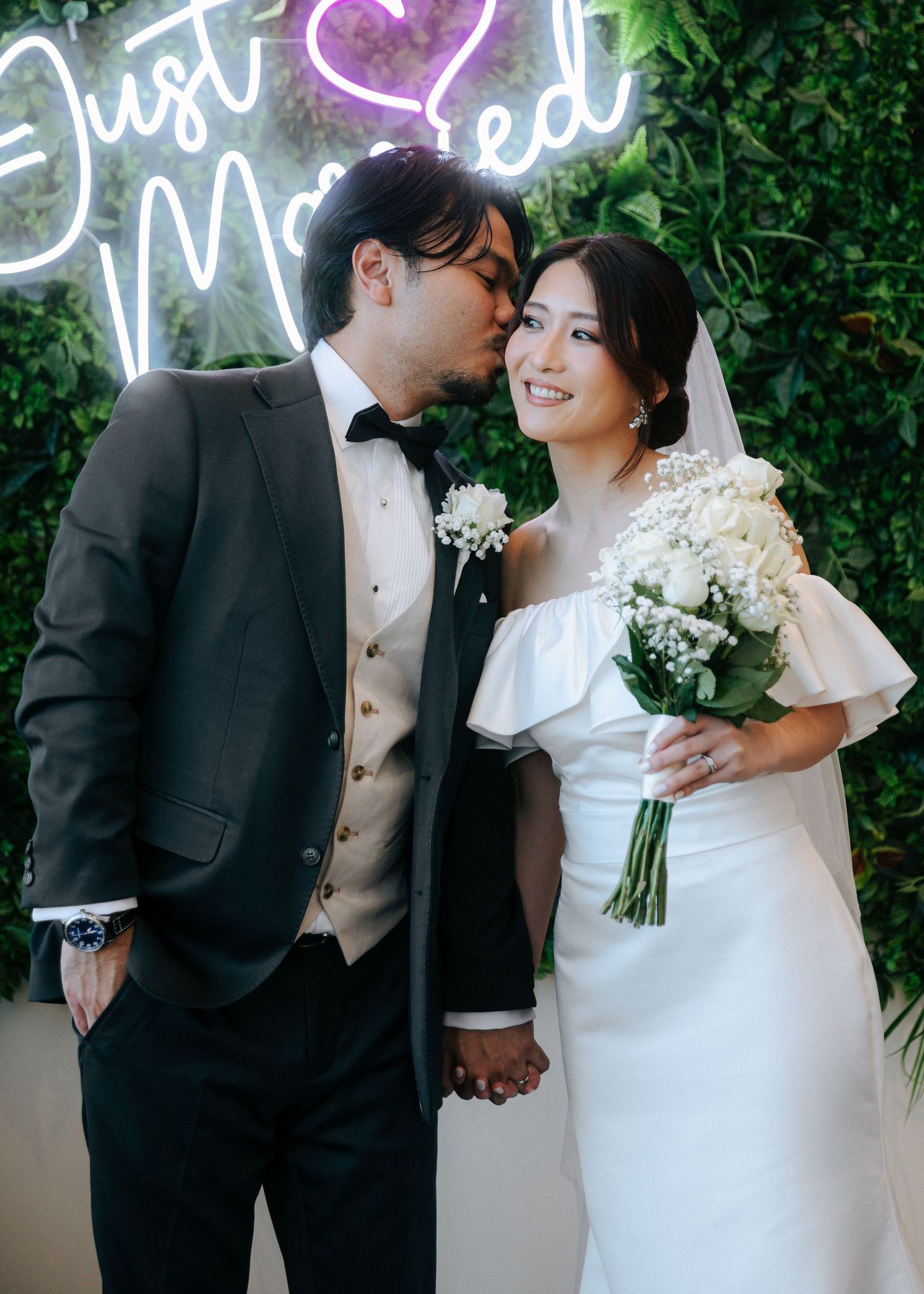 Bride and groom pose together under a Just Married neon sign, holding hands as groom kisses her cheek.