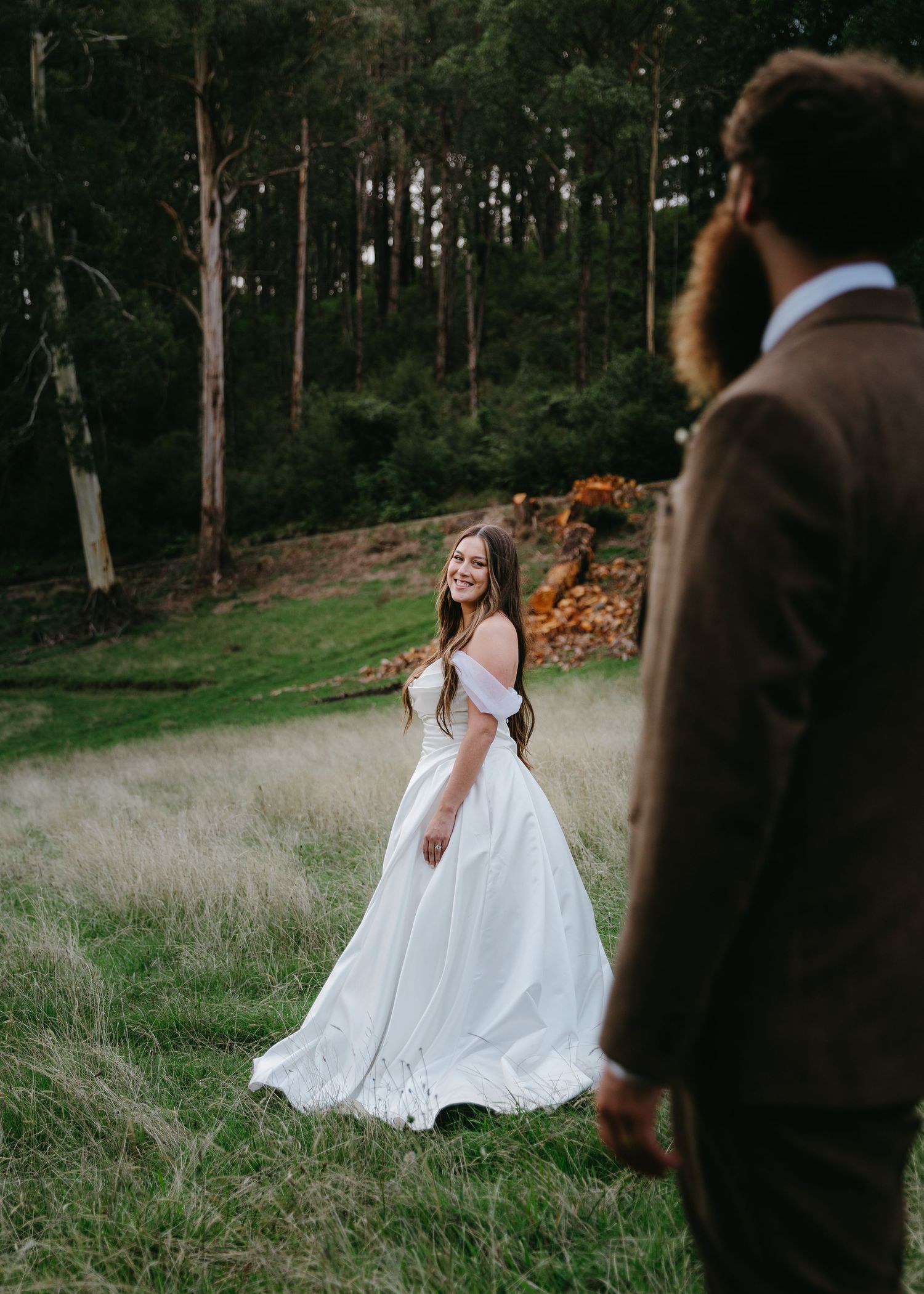 Smiling bride in white off-shoulder gown twirls in forest meadow.