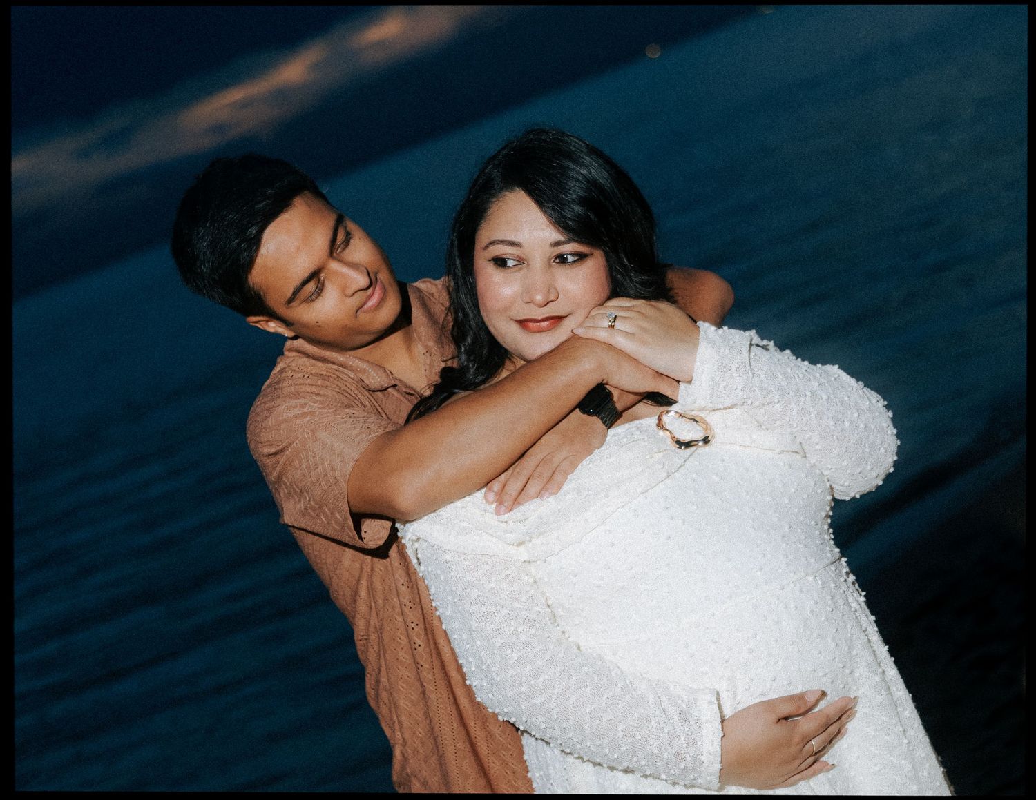 Couple embracing outdoors at dusk, man wrapping arms around pregnant woman in white dress.