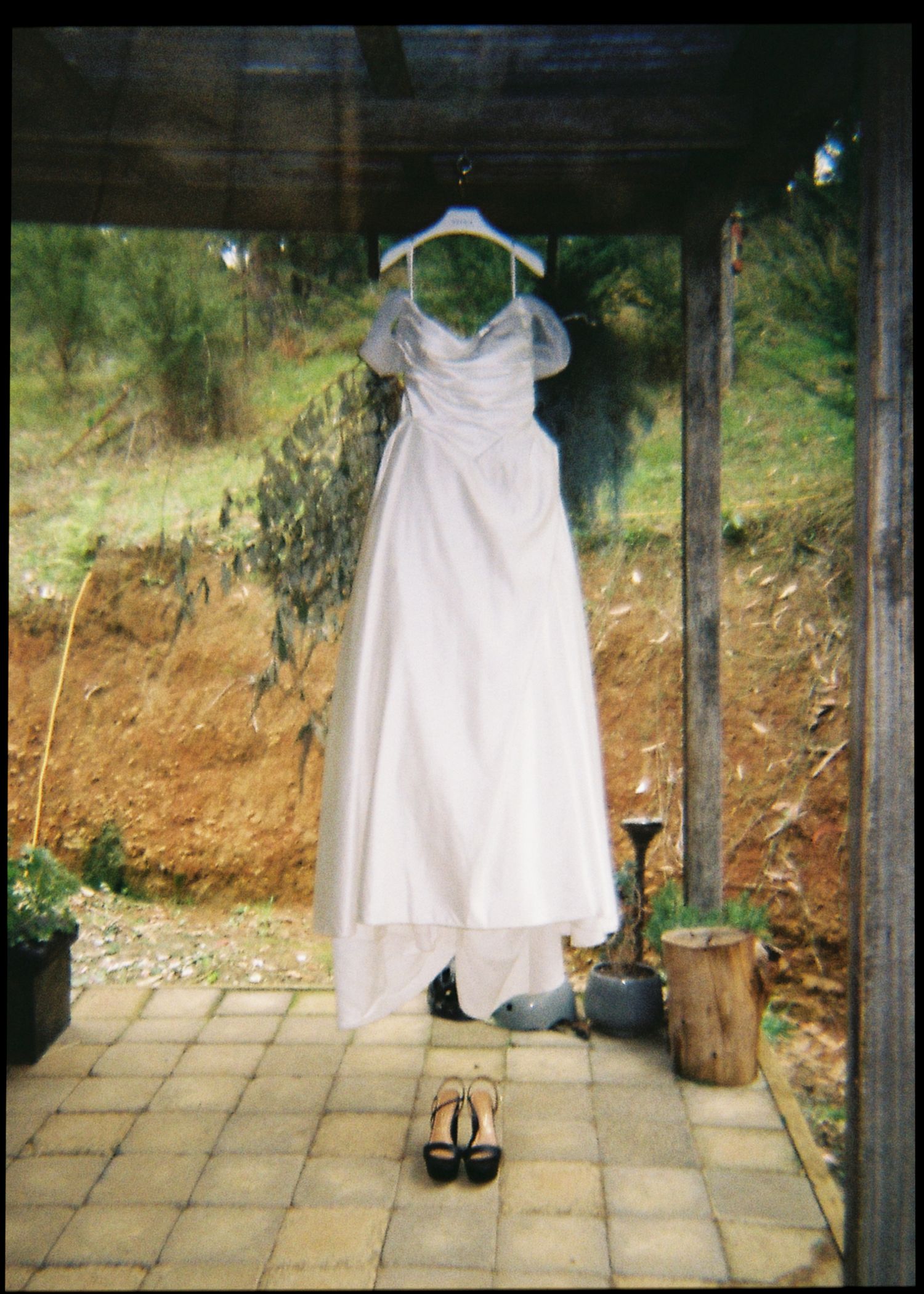 White wedding dress hanging on a hanger outdoors with black heels placed below on a tiled porch.