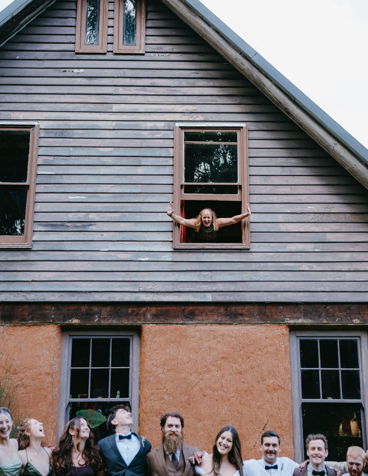 Wedding party posing outside a rustic barn, with a joyful guest leaning out of an upper window.