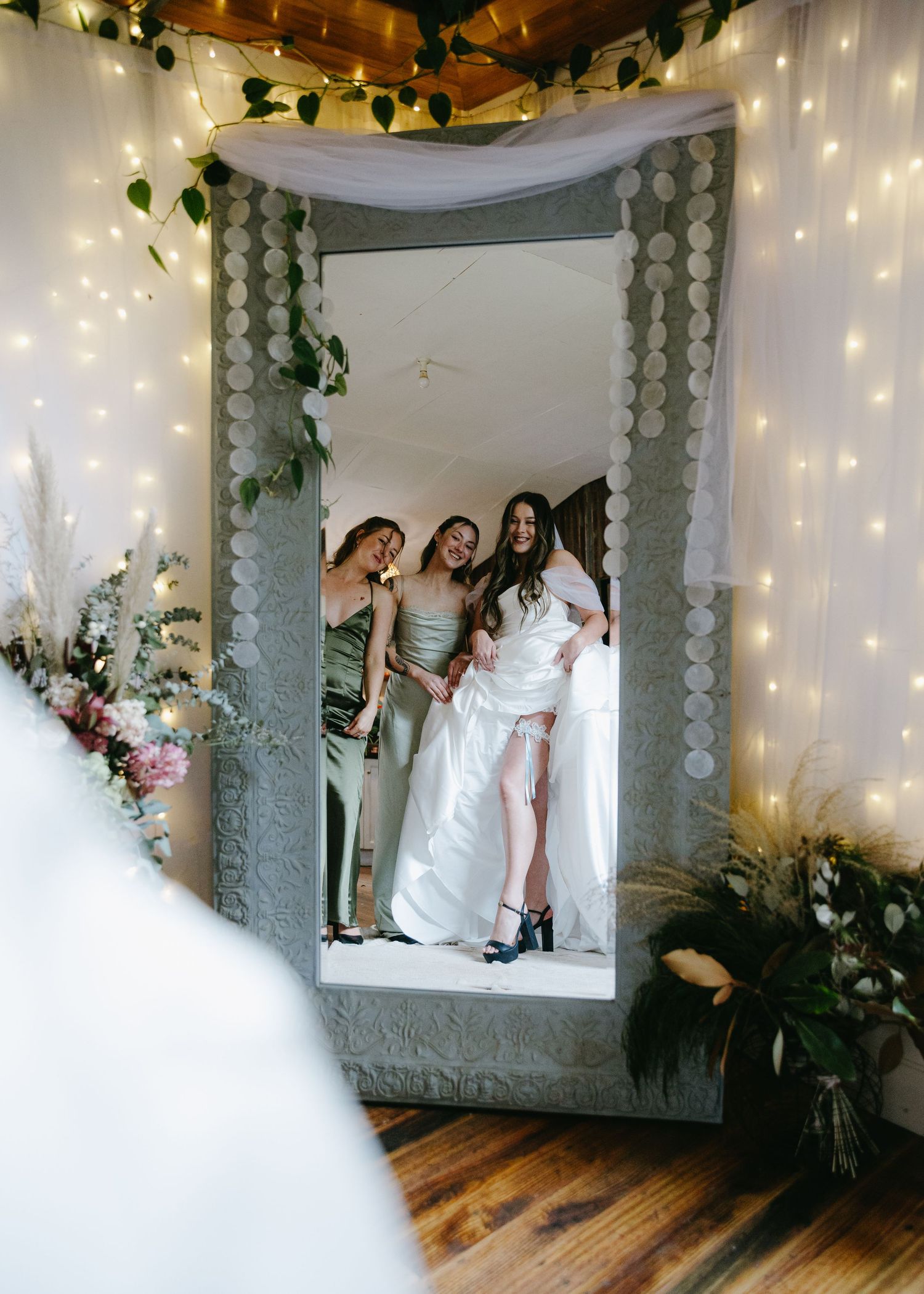 Bride and bridesmaids in sage green dresses laughing together reflected in a large mirror with fairy lights.