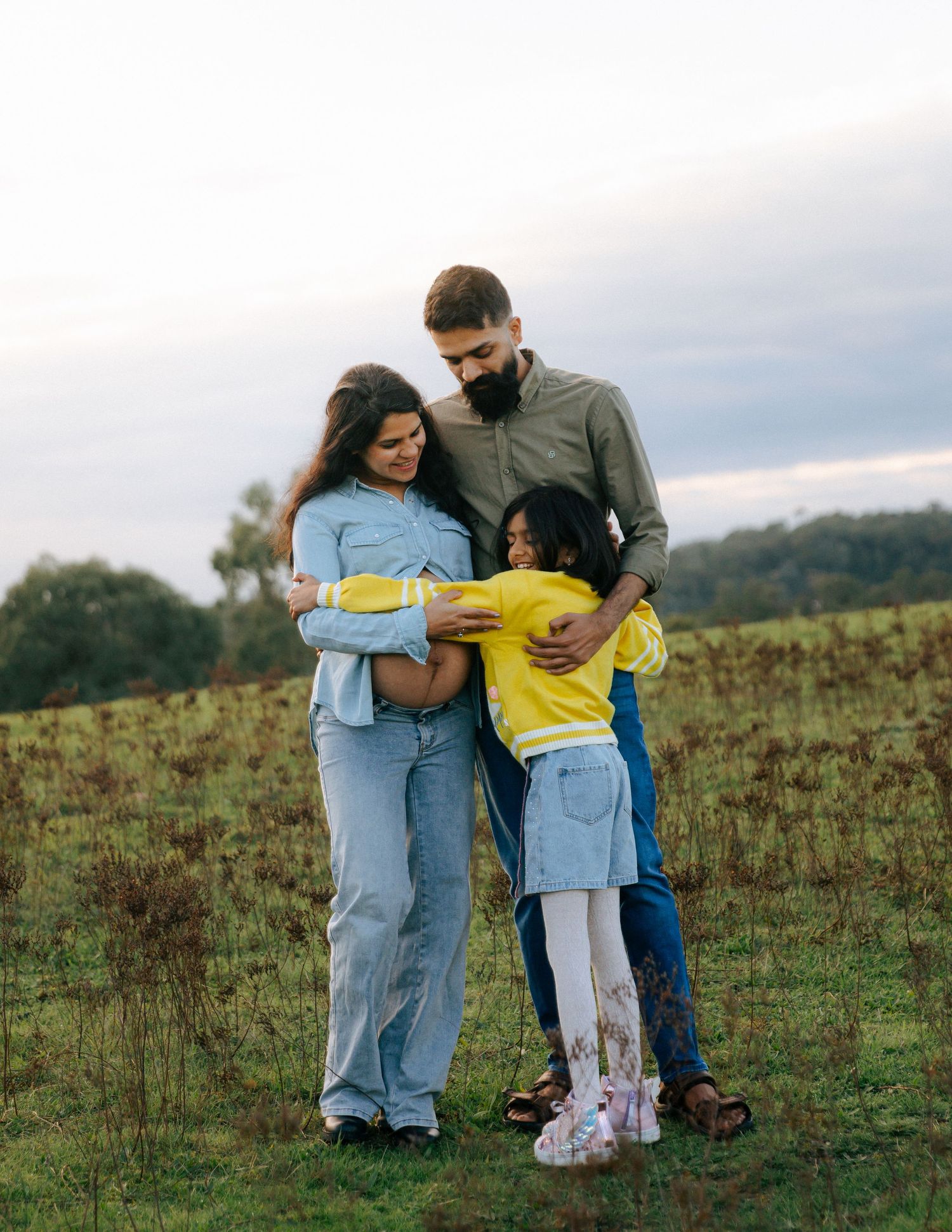 Family group hug with pregnant mother, father, and young daughter in field.