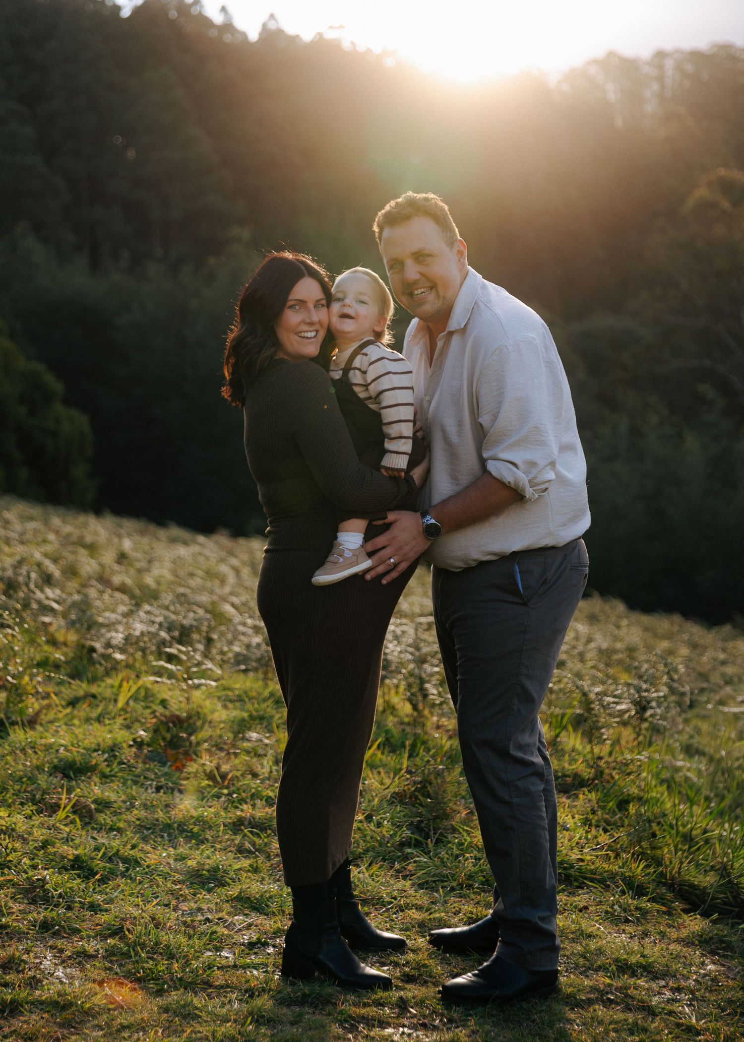 Happy family portrait at golden hour with parents holding baby in meadow.