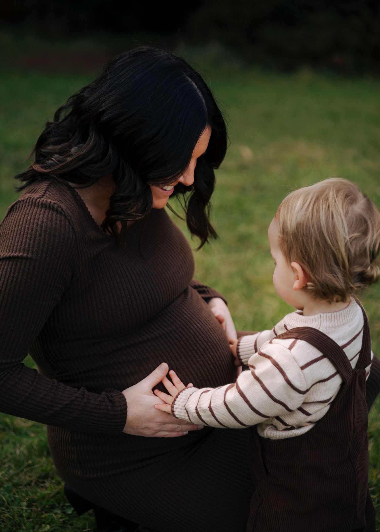 Pregnant woman in brown dress smiling at toddler touching her baby bump outdoors in green garden setting.