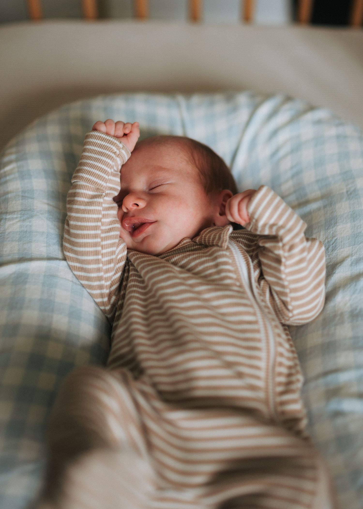 Sleeping newborn baby in striped onesie with arms raised in a cozy blue crib.
