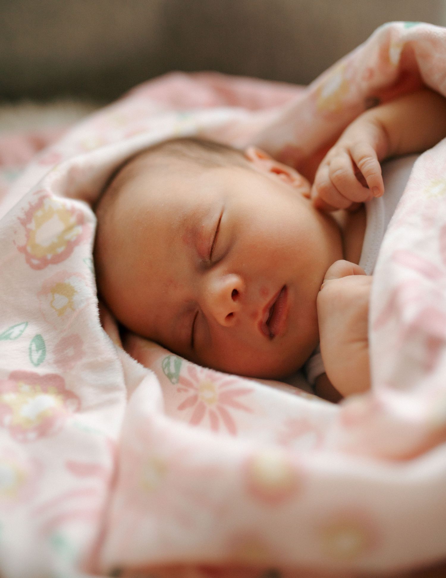 Sleeping newborn baby wrapped in soft pink floral blanket with tiny hands visible.