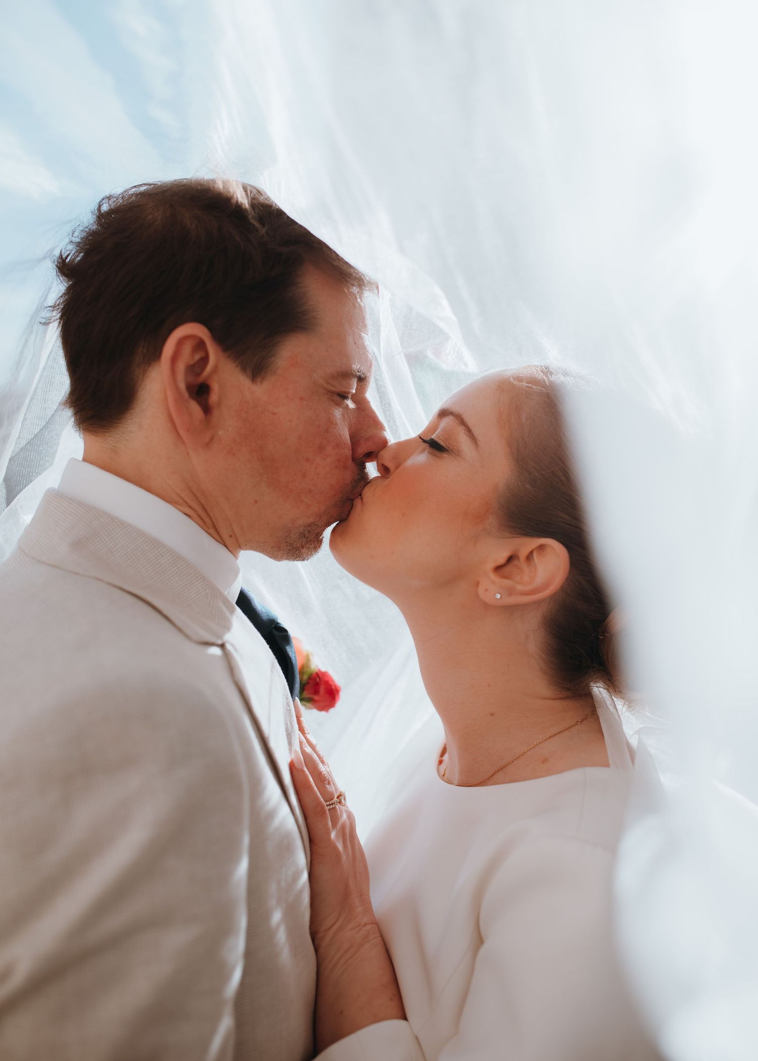 Tender wedding kiss captured under bridal veil with groom in beige suit and colorful boutonniere.