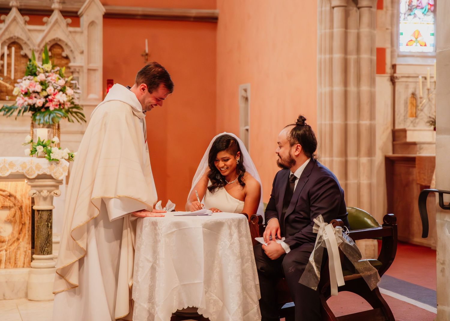 Bride signs the wedding register at a small table with the groom and officiant inside an elegant church.
