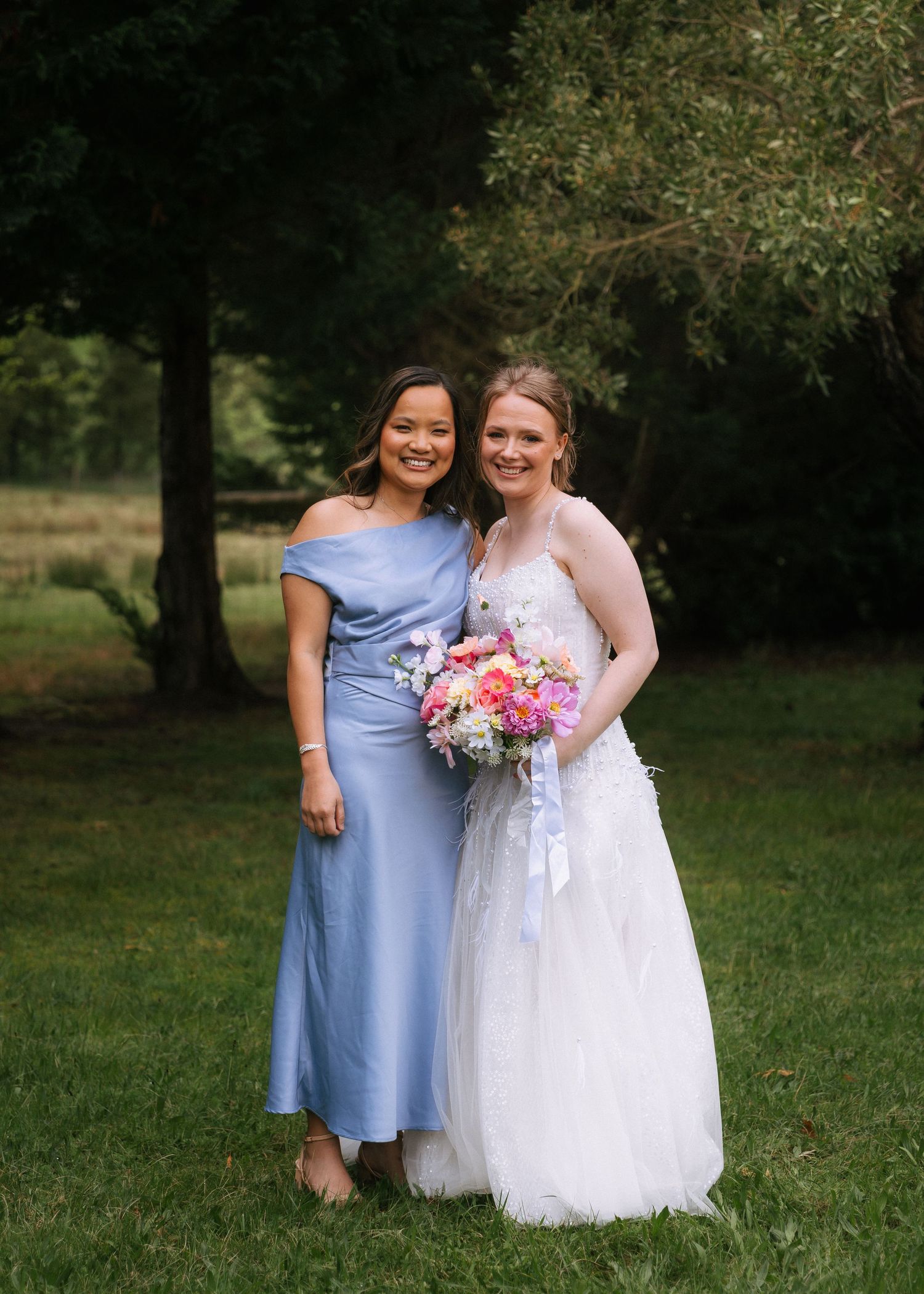 Bride in a white lace wedding gown holding a pink bouquet poses with a smiling bridesmaid in a blue dress on a grassy lawn.