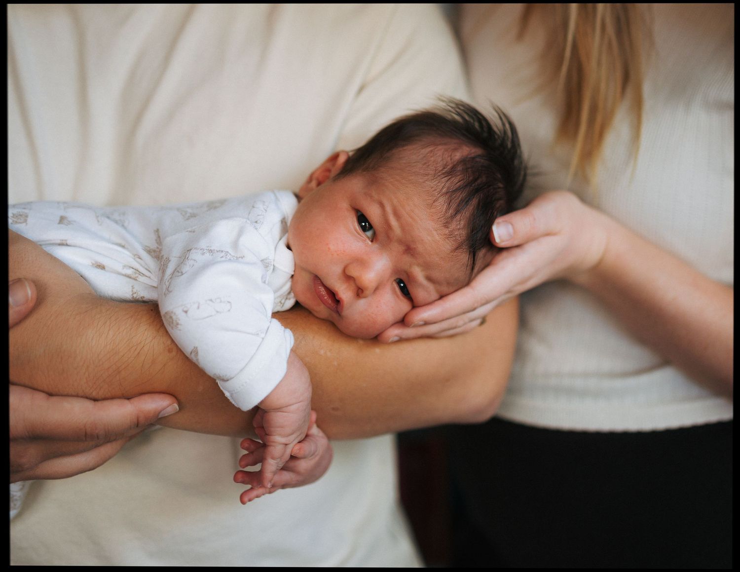 Newborn baby with dark hair rests on a parent's forearm while another adult gently cradles the infant's head in a warm, tender moment.