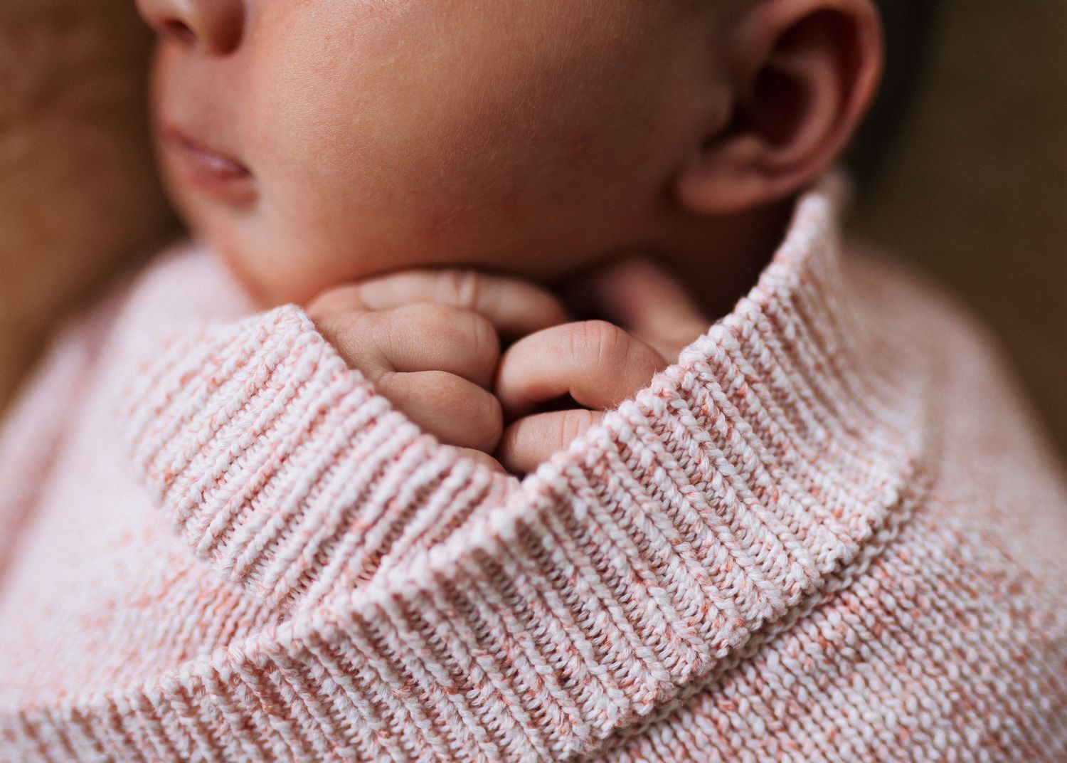 Close-up of a newborn baby's tiny folded hands tucked under the chin, wrapped in a soft pink knit blanket.