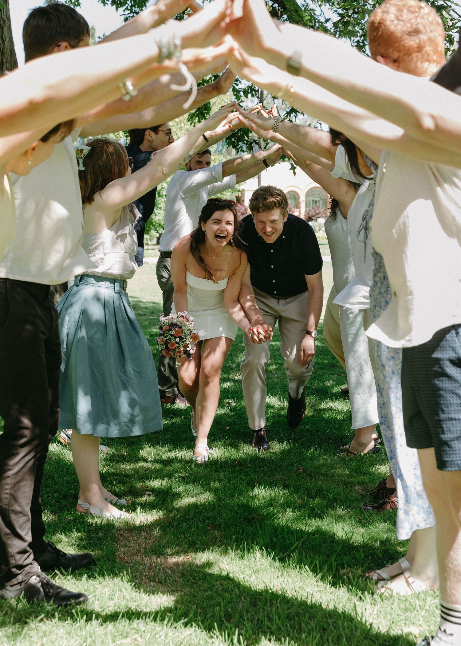 Wedding guests forming an arch with their arms as newlyweds run through laughing on sunny day.
