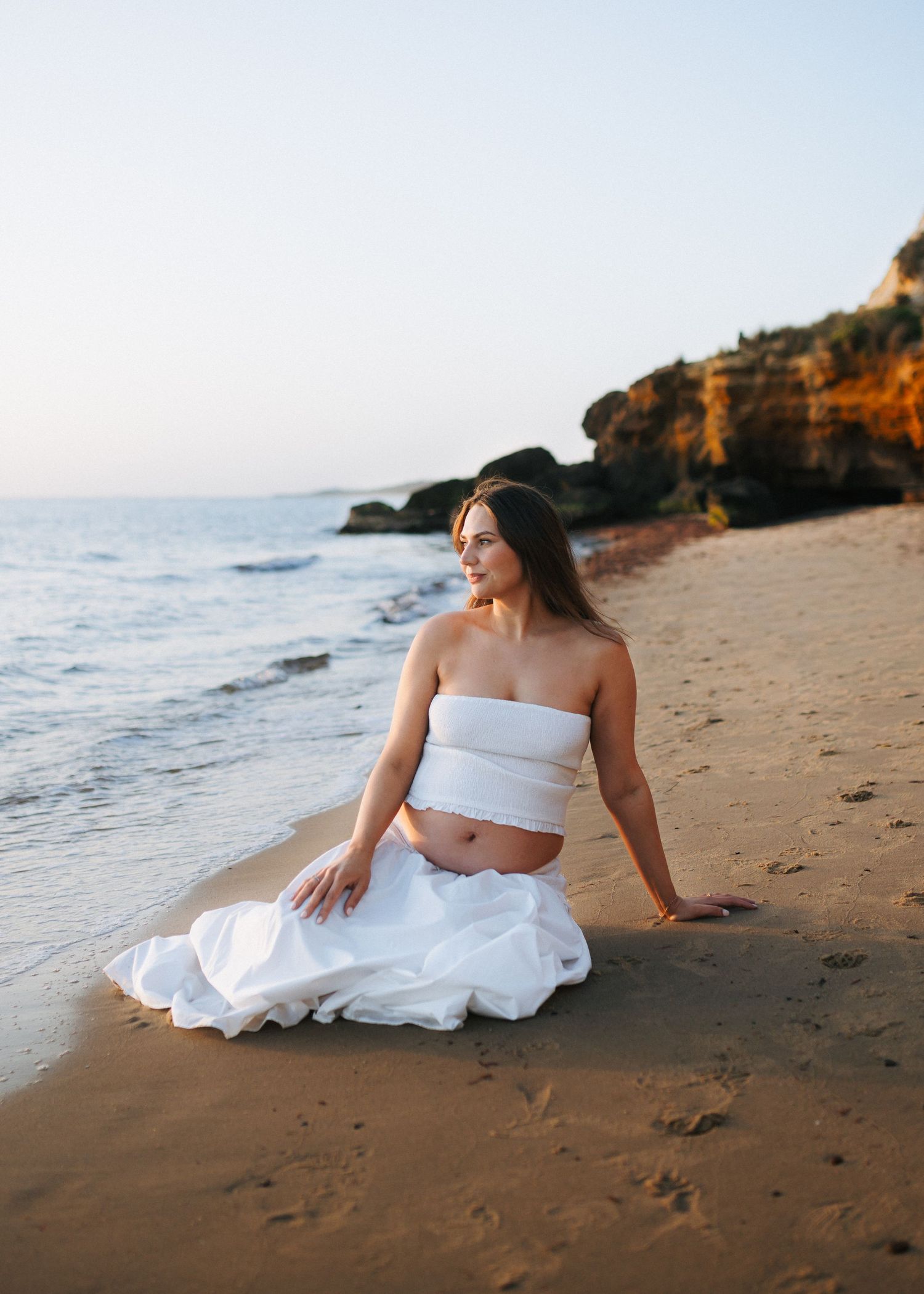 Pregnant woman in white bandeau top and skirt sitting gracefully on sandy beach at sunset with rocky cliffs in background.
