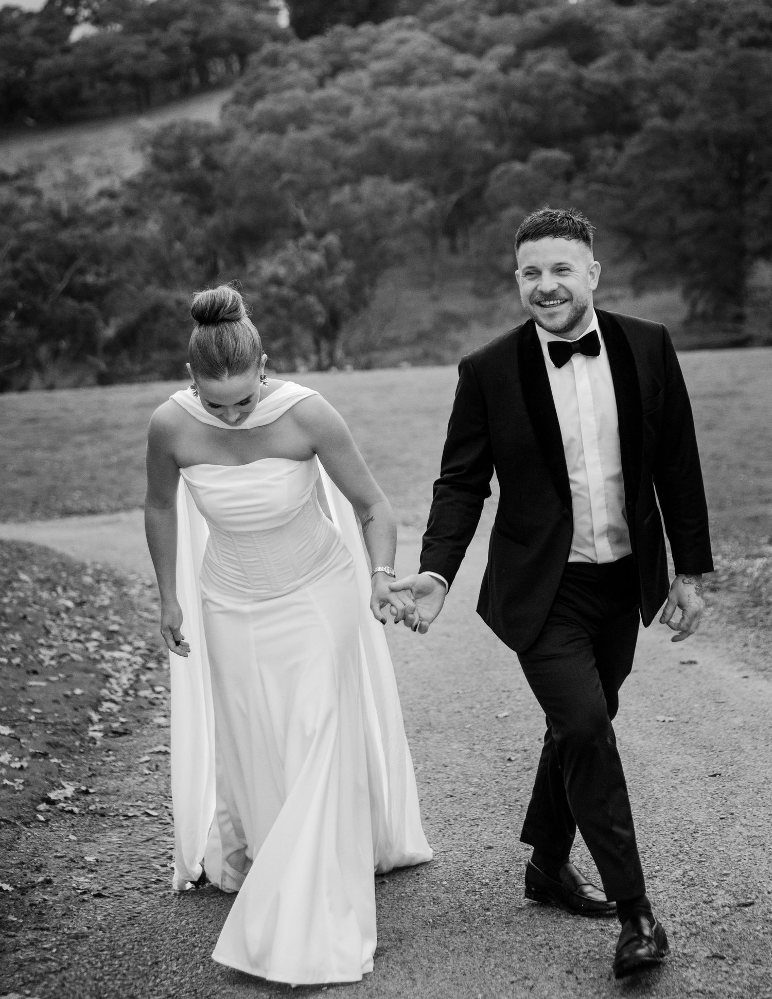 Black and white photo of bride and groom walking hand in hand along country road.
