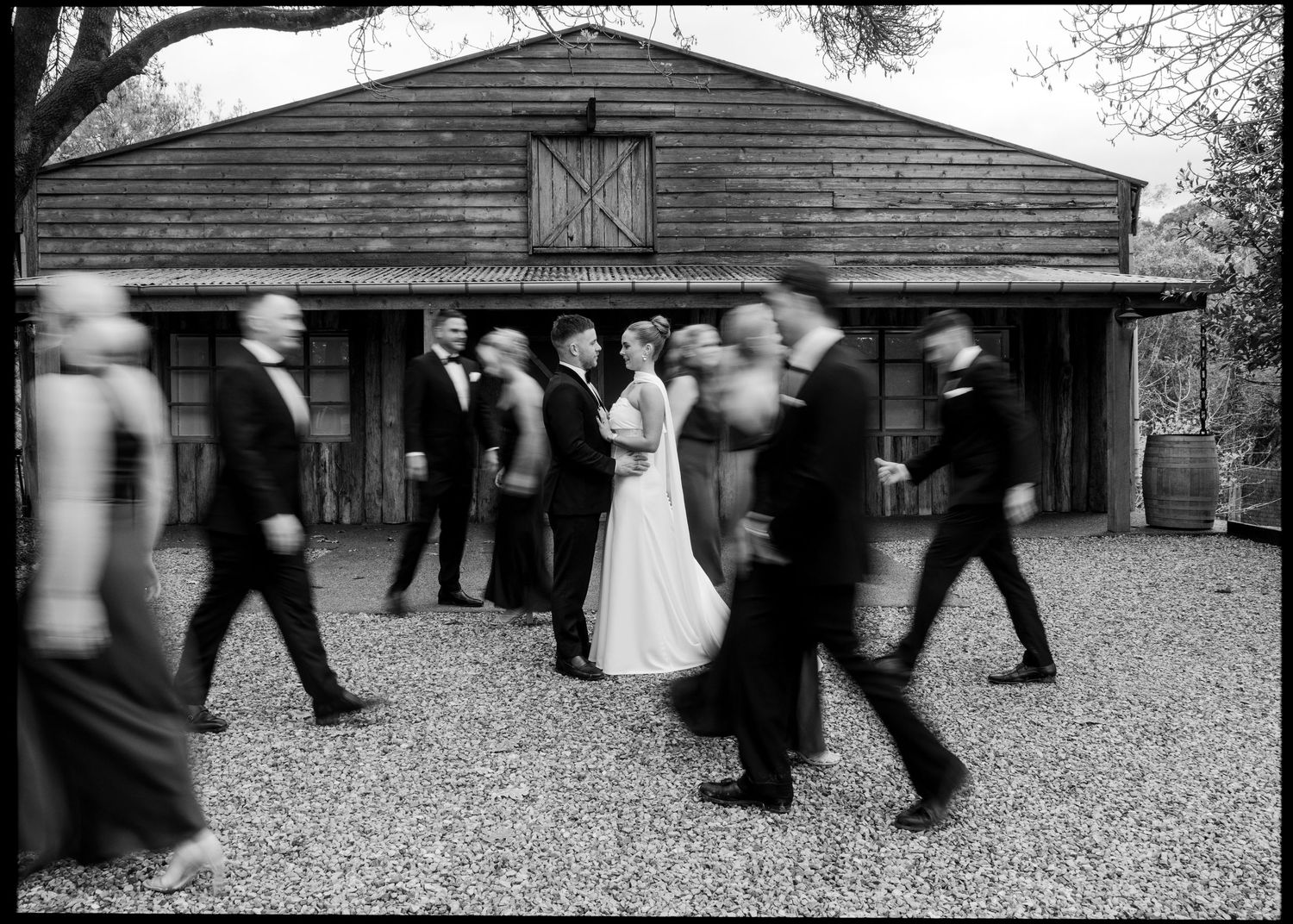 Black and white wedding photo of bride and groom standing still as blurred wedding party walks past a rustic barn.