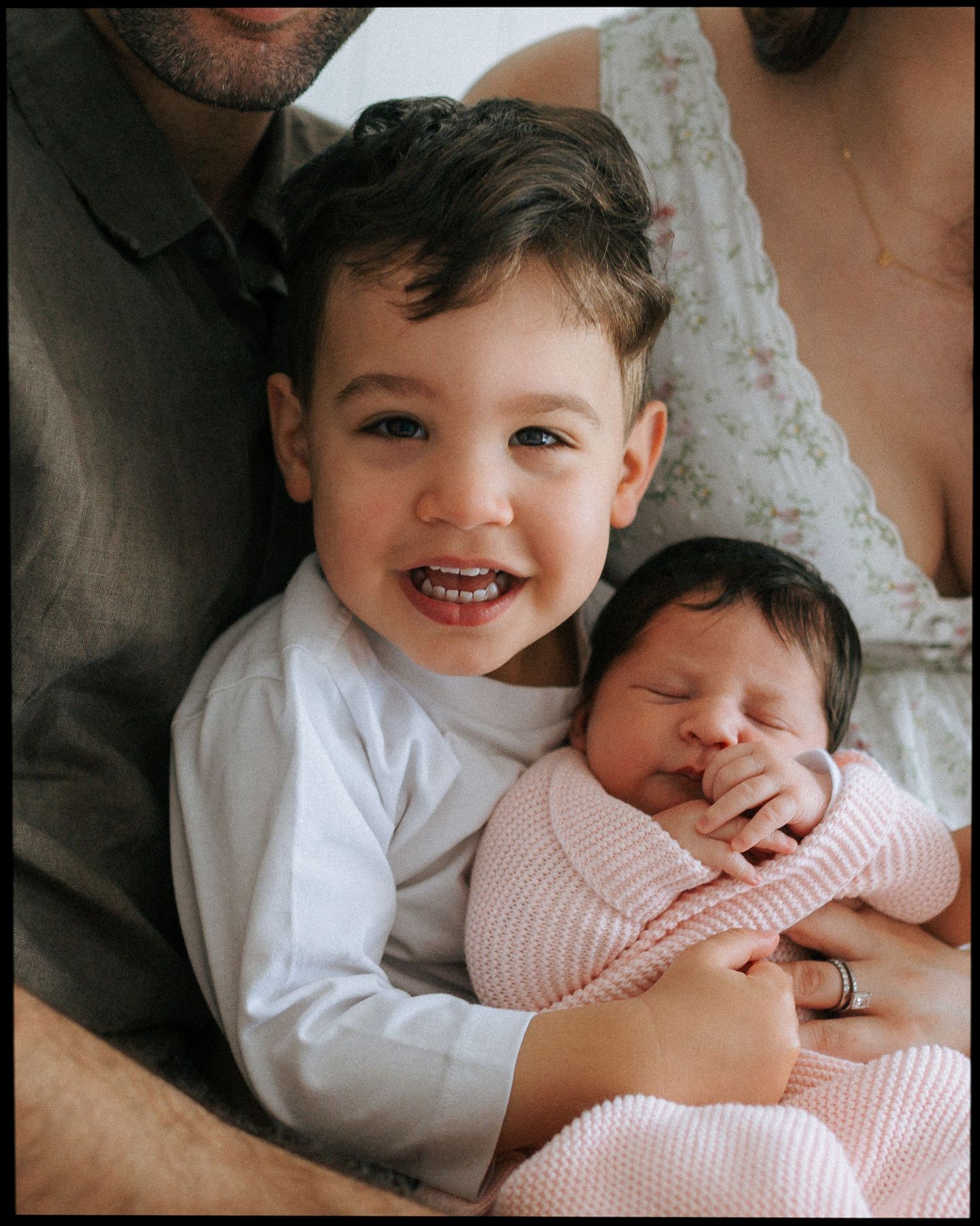 Adorable smiling toddler boy posing with sleeping newborn sibling wrapped in pink swaddle blanket.