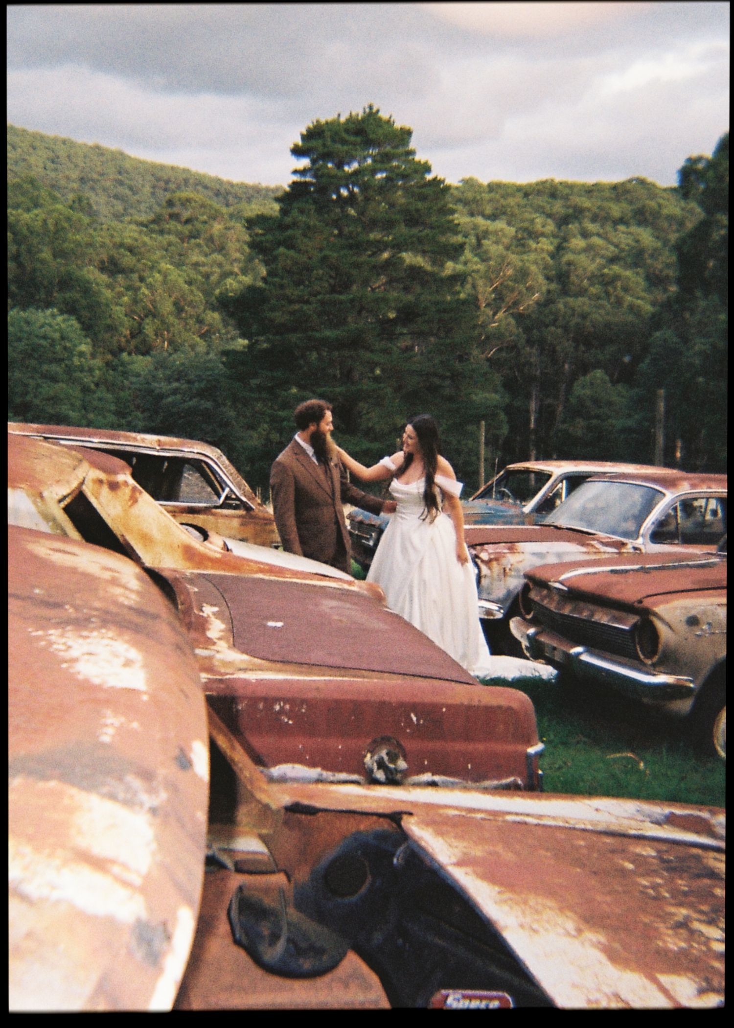 Bride in white gown and groom in suit share a tender moment among rusty vintage cars in a junkyard.