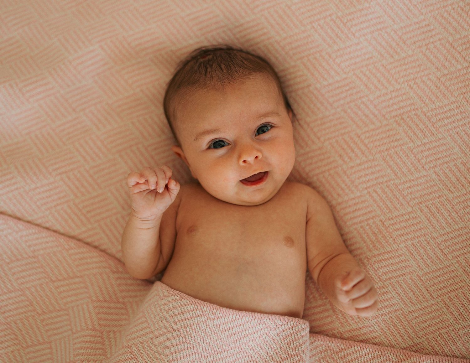 Adorable newborn baby lying on a pink textured blanket smiling upward.