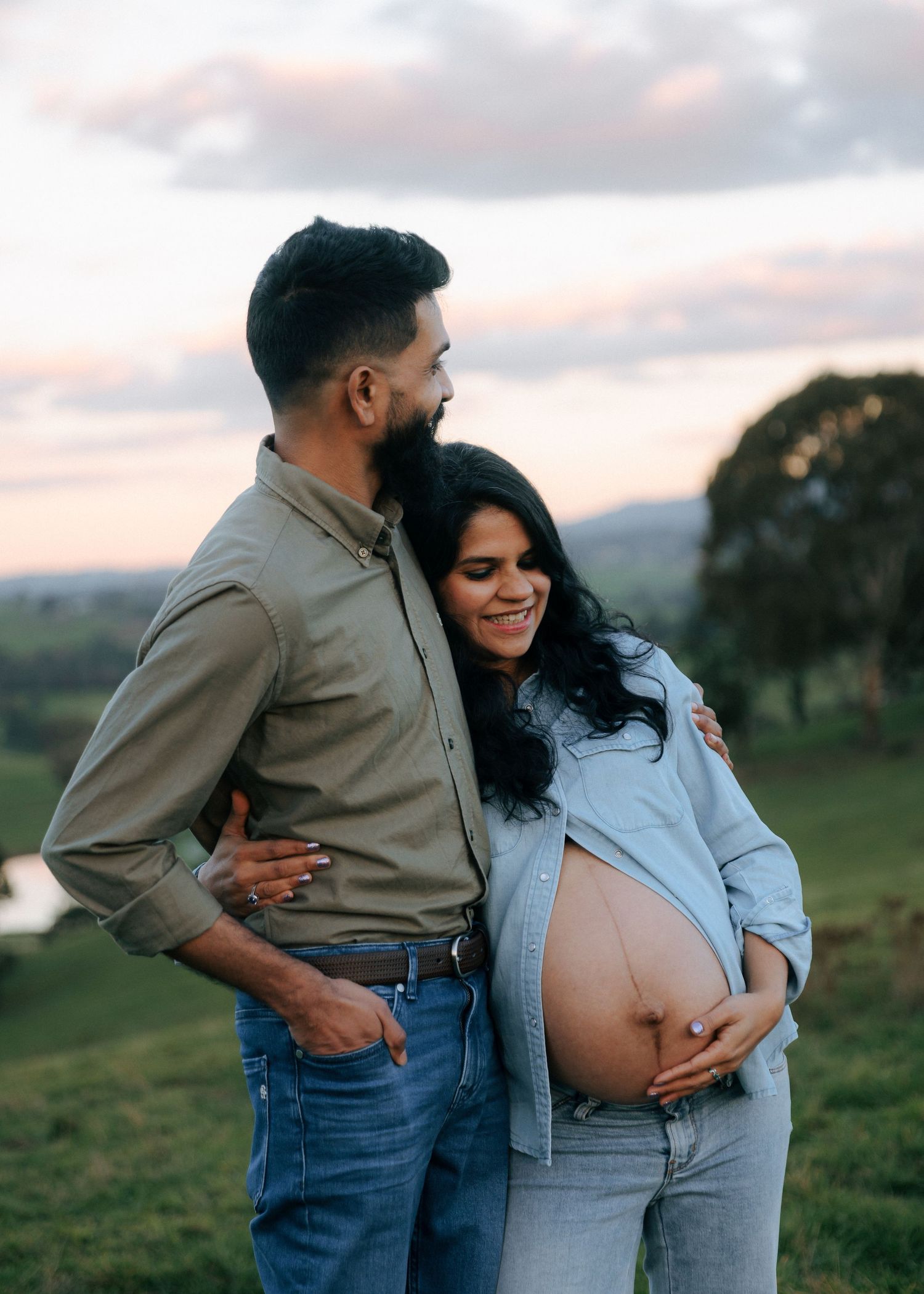 Smiling pregnant woman and partner embracing on a green hillside at sunset, cradling her exposed baby bump.