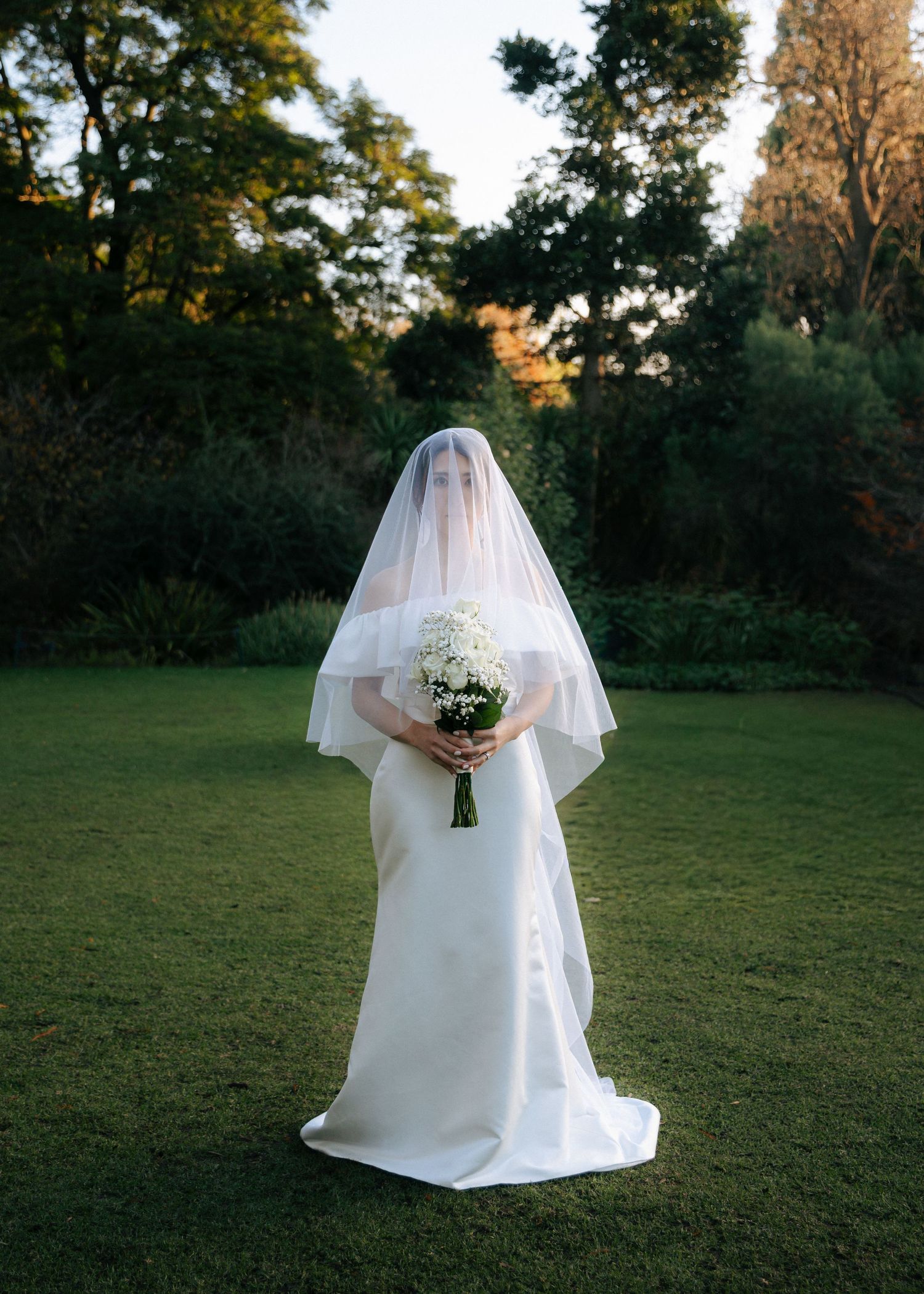 Bride in white satin gown and veil stands on green lawn holding a white bouquet, veil covering her face.