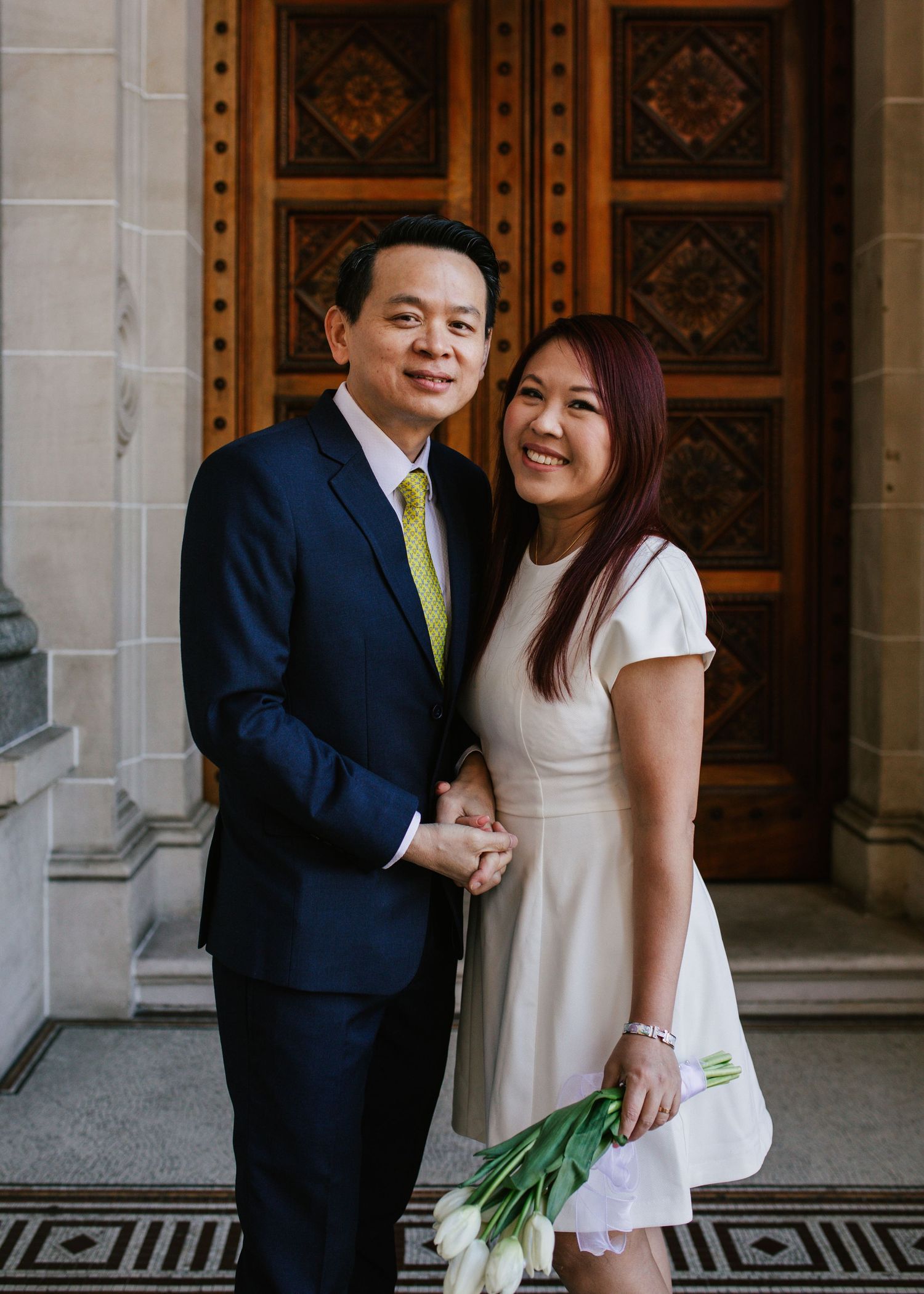 Smiling Asian couple posing with white tulips before ornate carved wooden doors at registry.