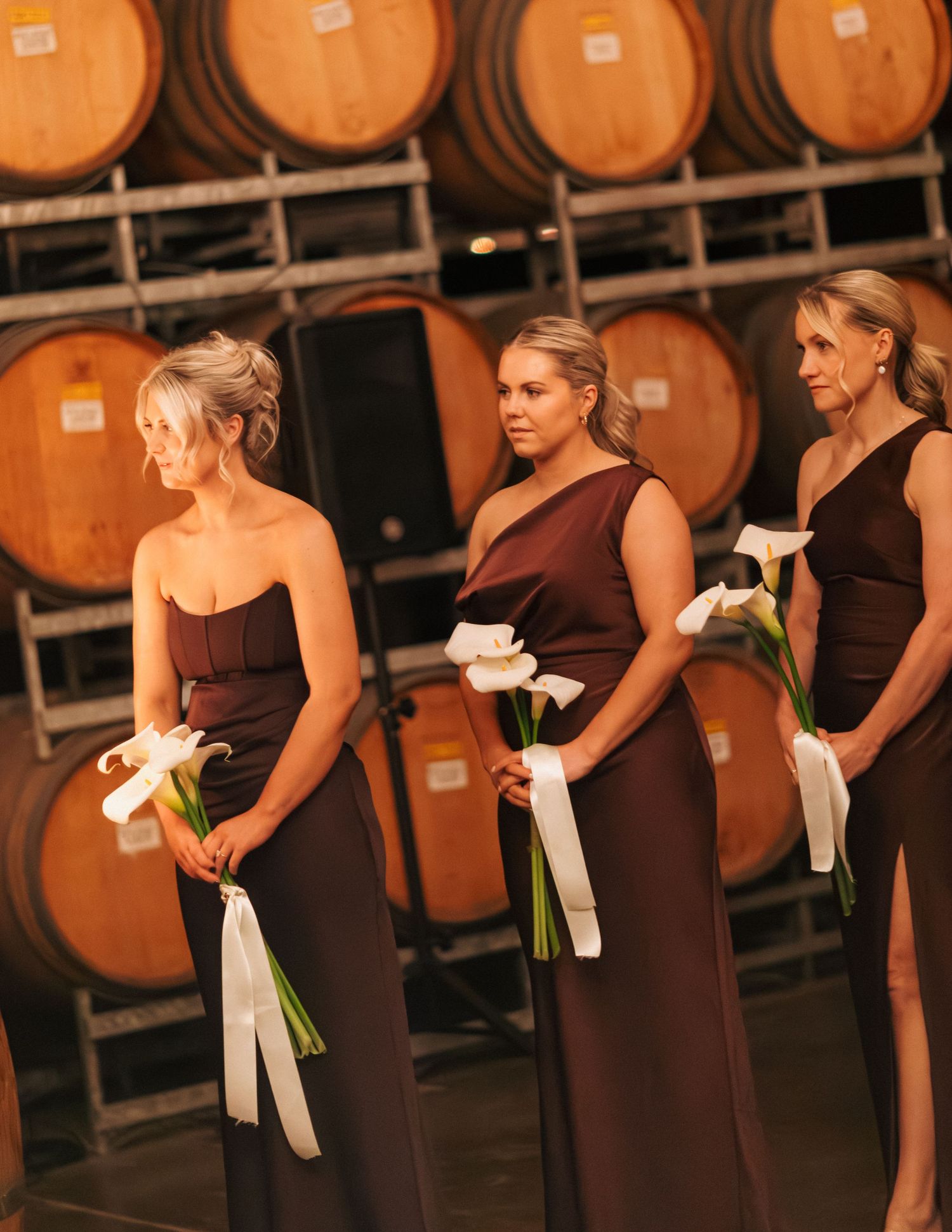 Three bridesmaids in dark brown gowns holding white calla lily bouquets in front of wine barrels.