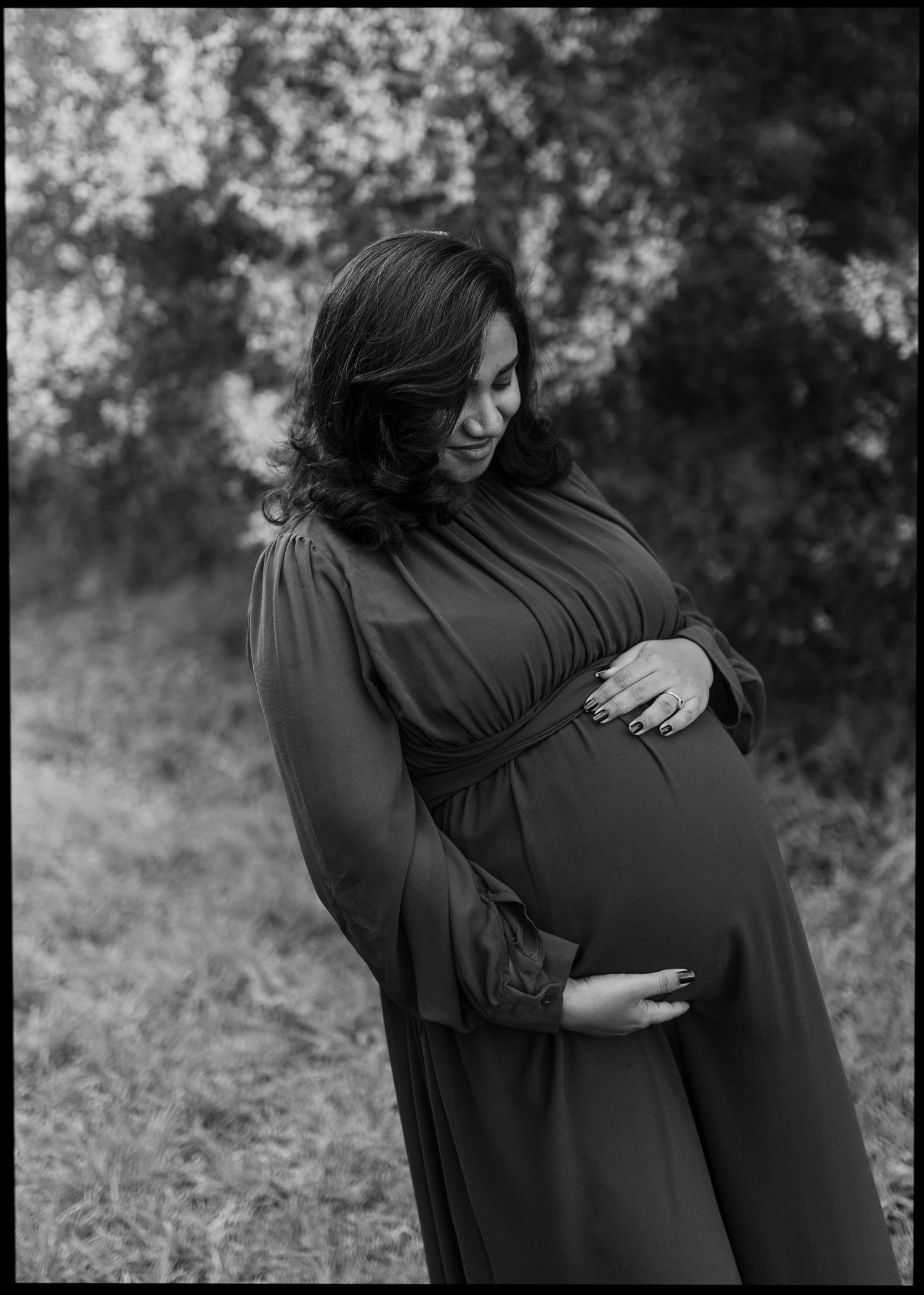 Black and white maternity portrait of a pregnant woman in a flowing long-sleeve dress cradling her belly outdoors among blurred trees.