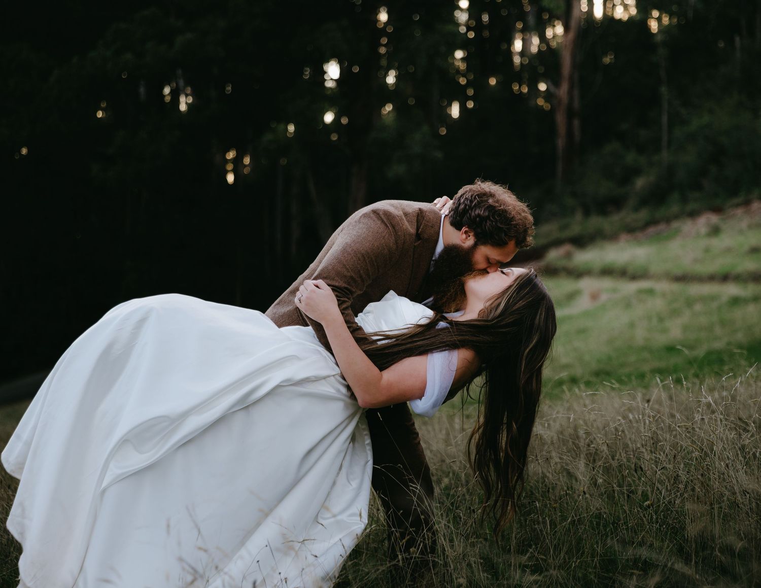 Groom dips bride in dramatic kiss in dark forest meadow setting.