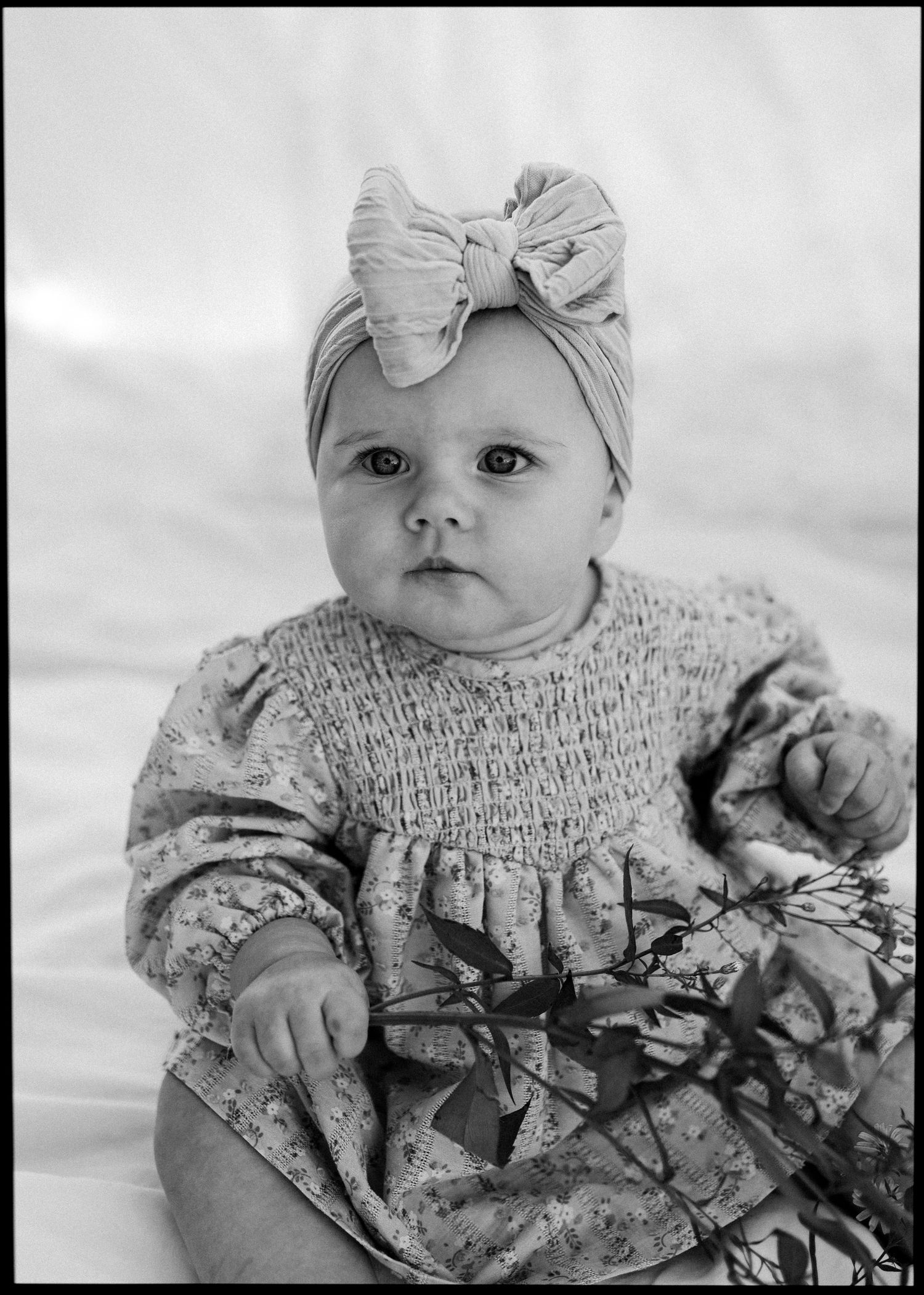Black and white portrait of baby girl in floral dress and large bow headband.