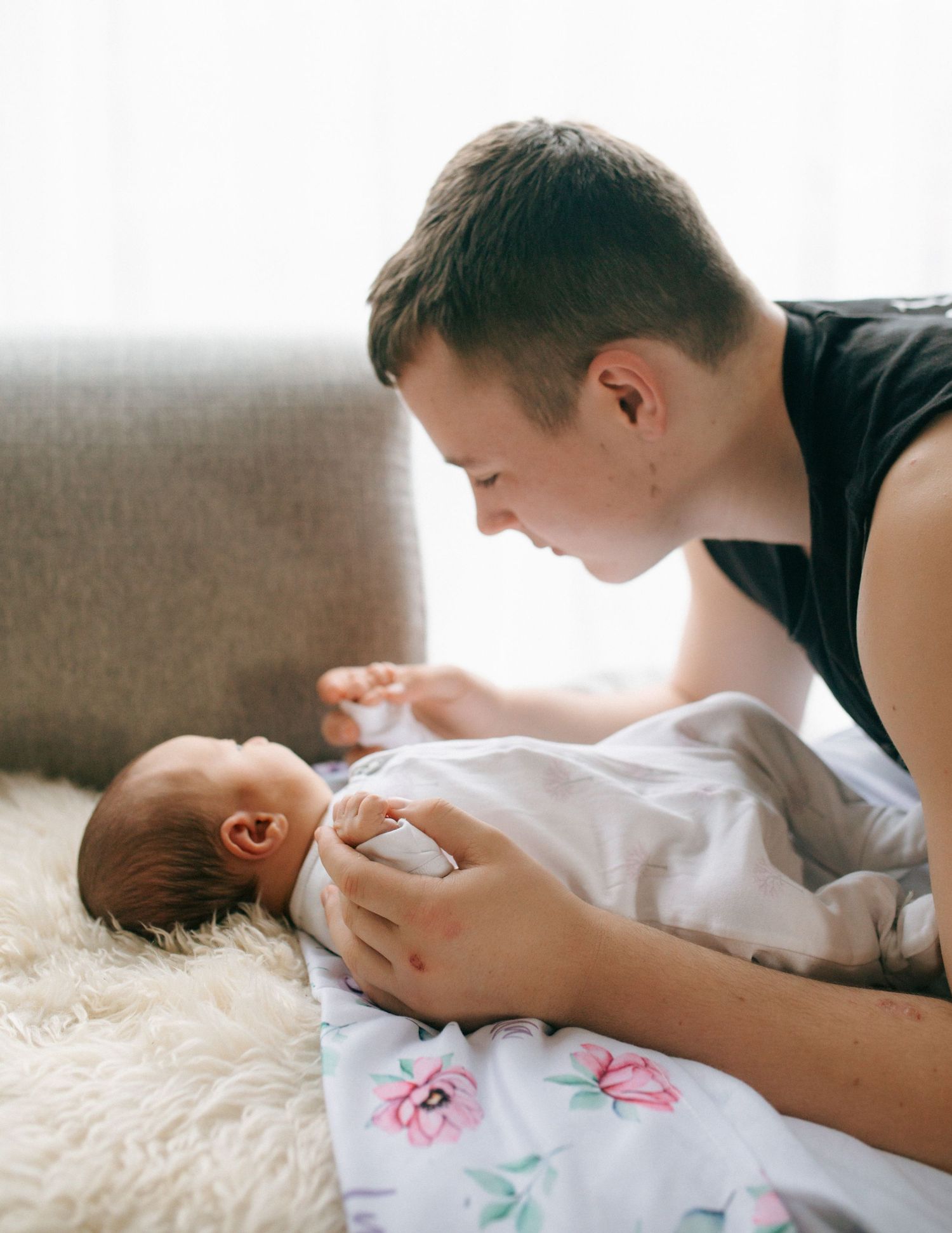 Father leaning over newborn baby swaddled in floral blanket on fluffy white rug.