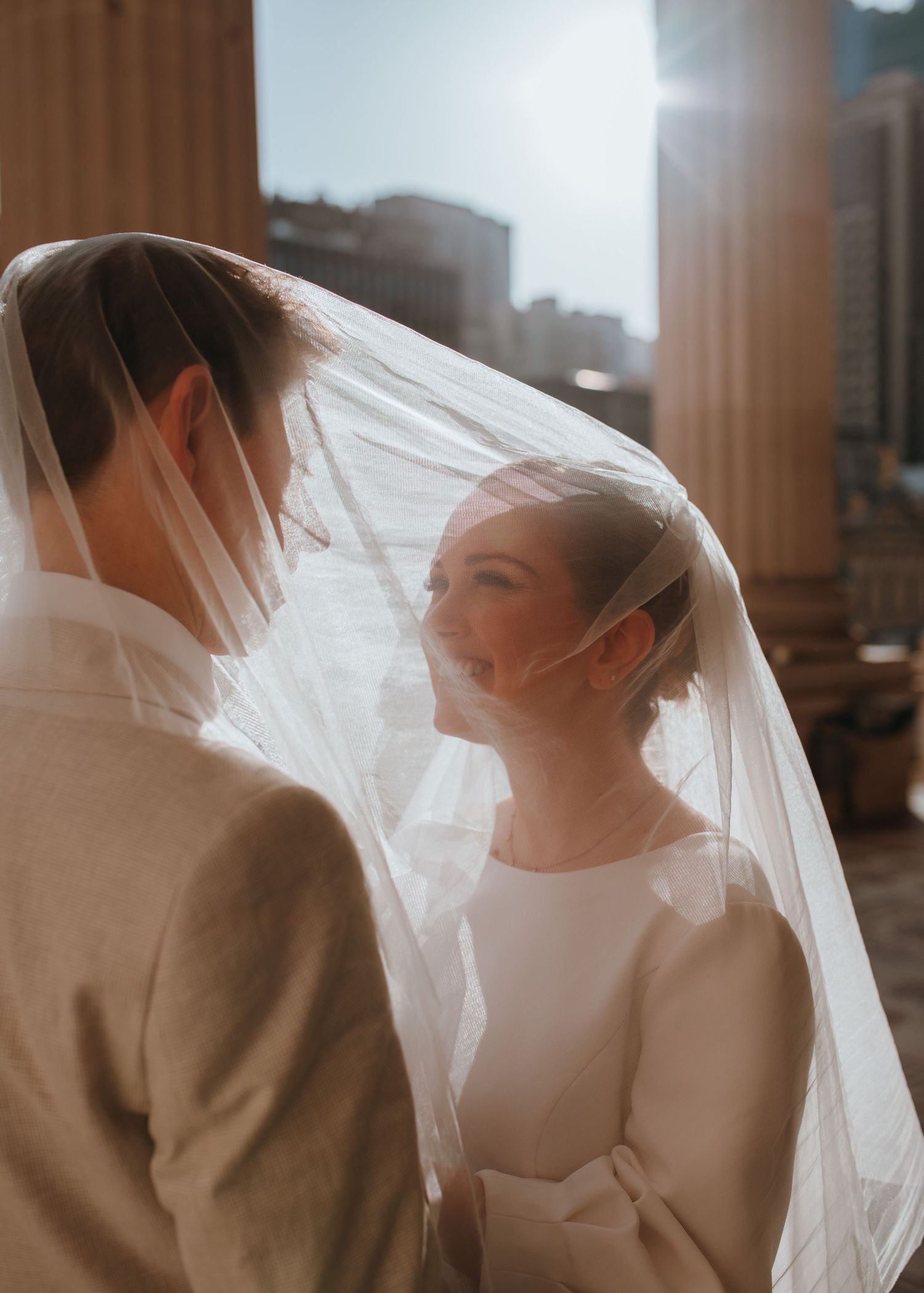 Romantic moment of bride and groom under flowing veil with sunlit city building in background.