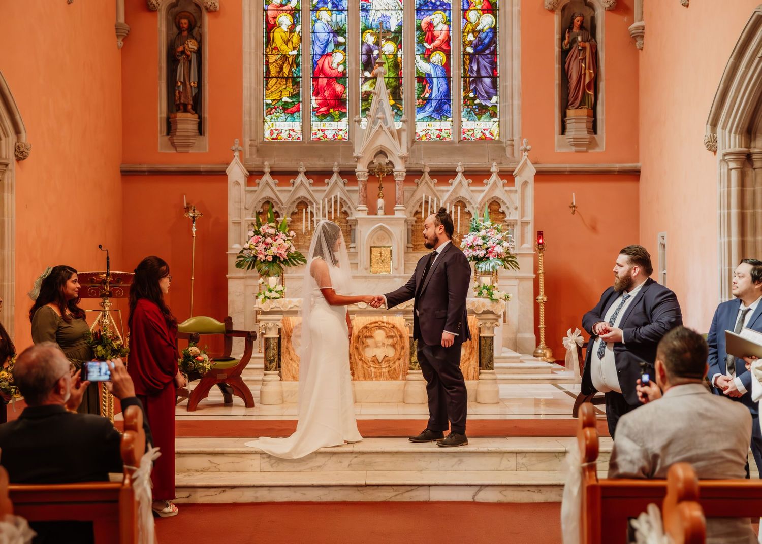 Bride and groom exchange vows at the altar of a stunning church with colorful stained glass windows.