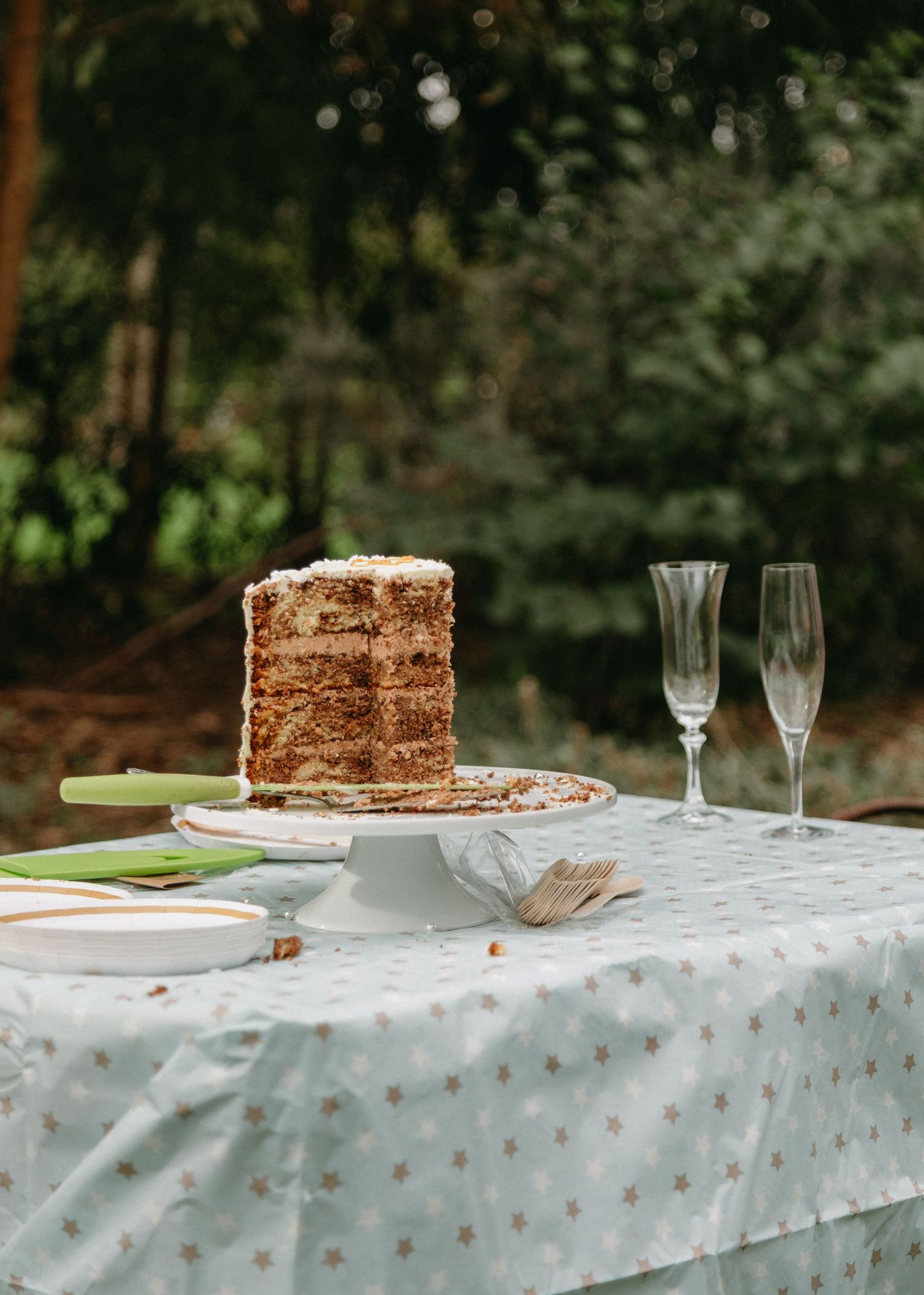 Tall layered wedding cake on white stand with champagne flutes on star-print tablecloth outdoors.