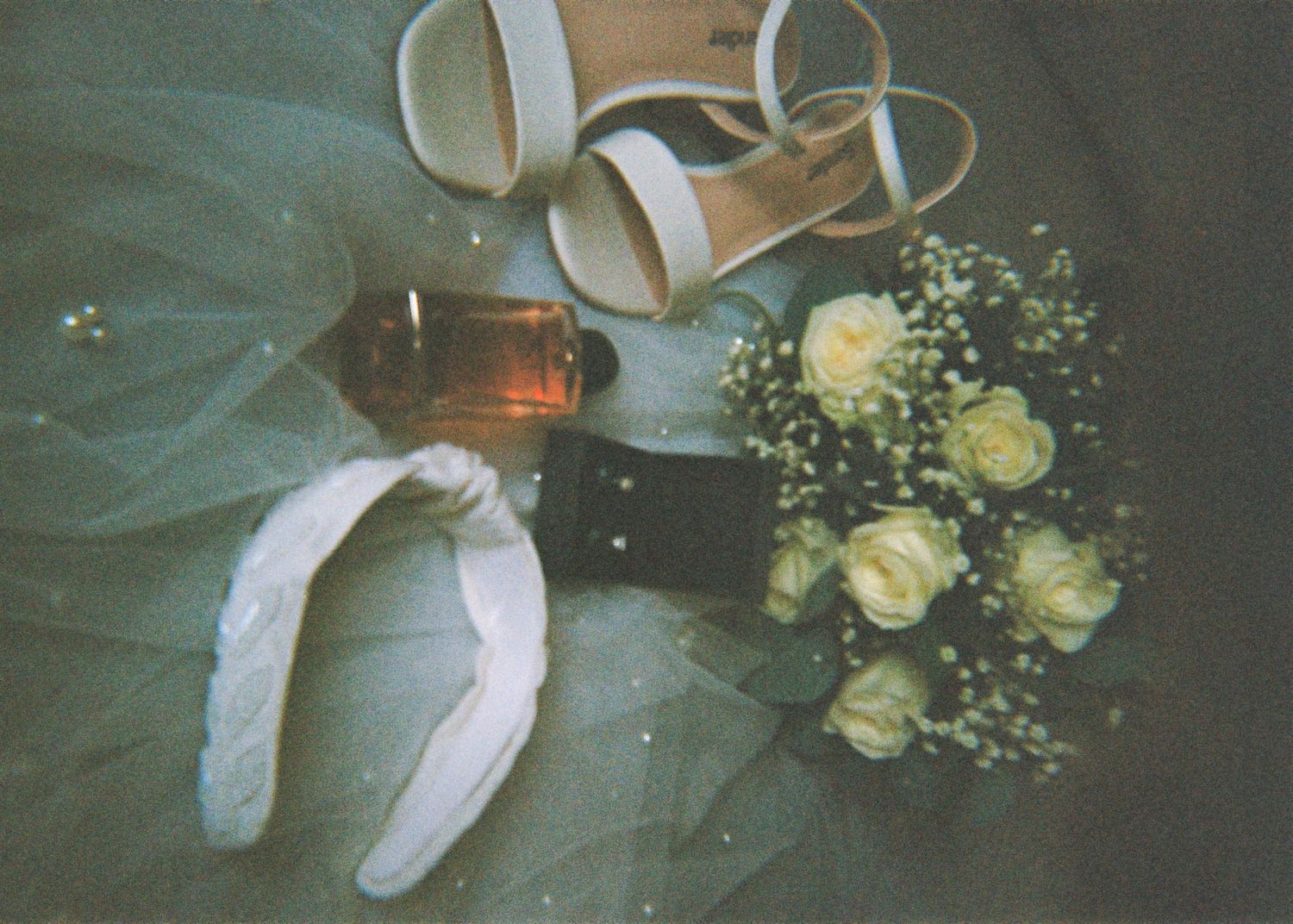 Flat lay of bridal accessories featuring white rose bouquet, strappy heels, perfume bottle, and hair bow on blue tulle.