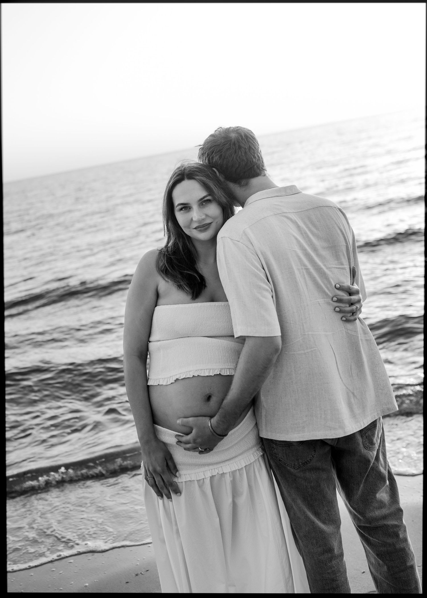Black and white beach maternity photo of pregnant woman in white dress with partner kissing her cheek by the ocean.